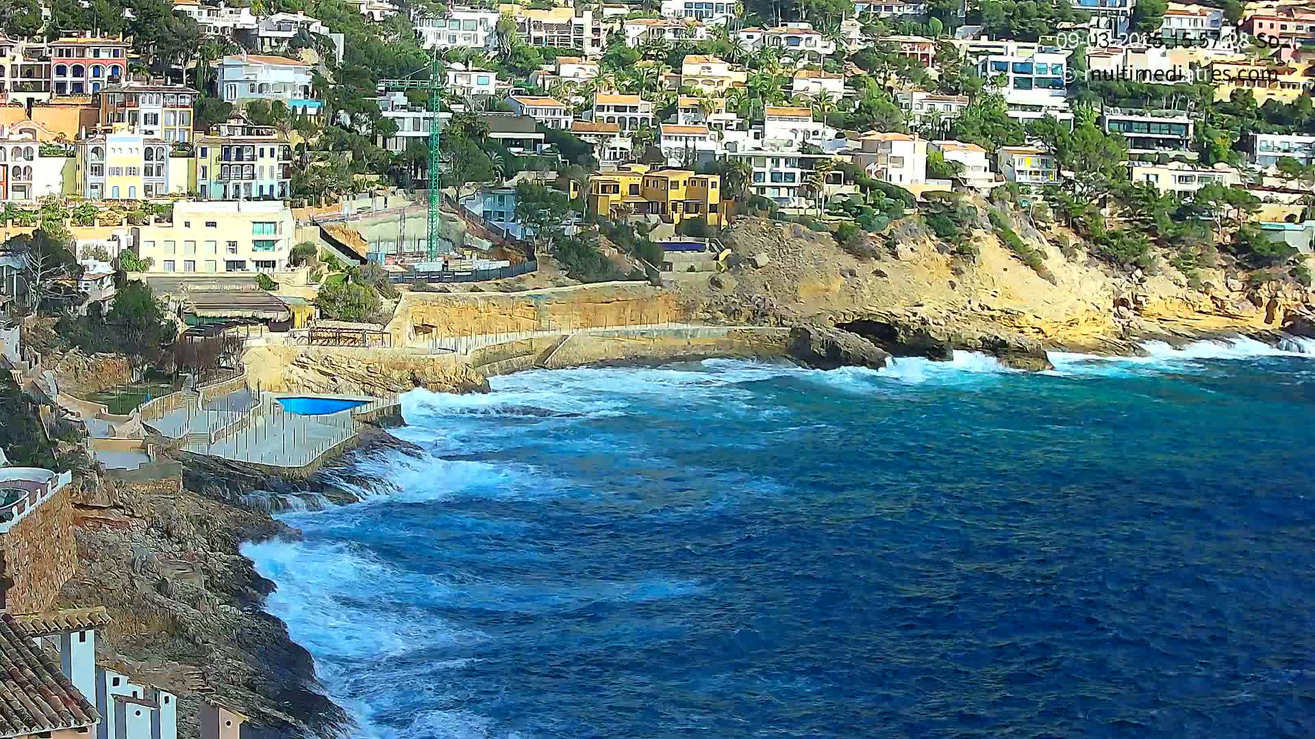 A rocky coastline with houses built into a hillside overlooks a wavy, blue ocean.