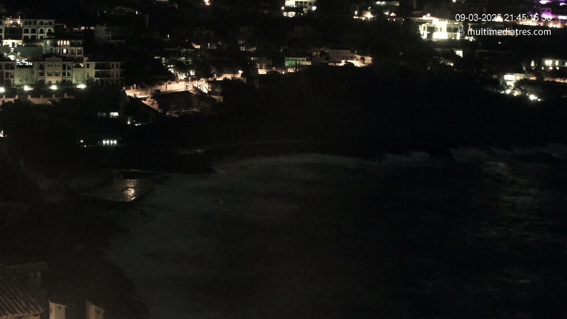 A nighttime coastal scene shows a dark ocean with some small waves, a dark hillside with dimly lit buildings, and some lights visible on the buildings themselves.