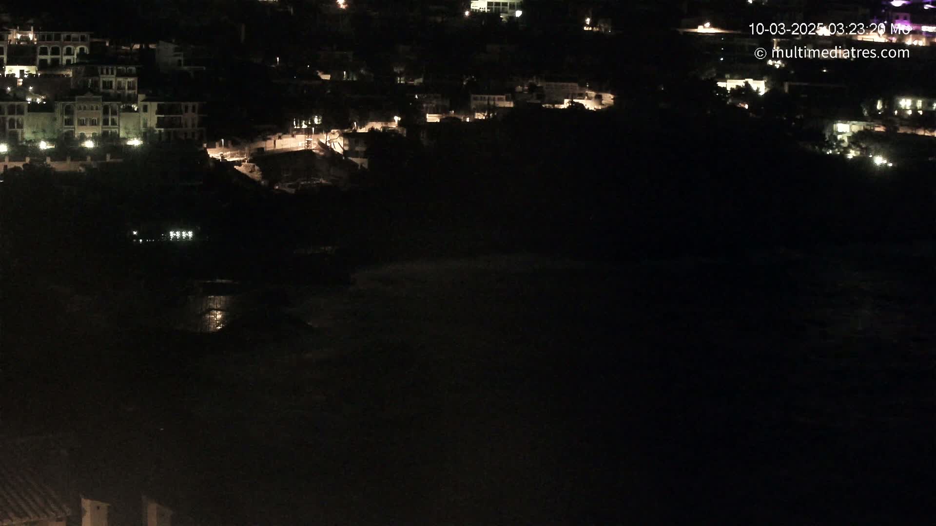A nighttime, high-angle view shows a dark body of water below a hillside of dimly lit buildings.