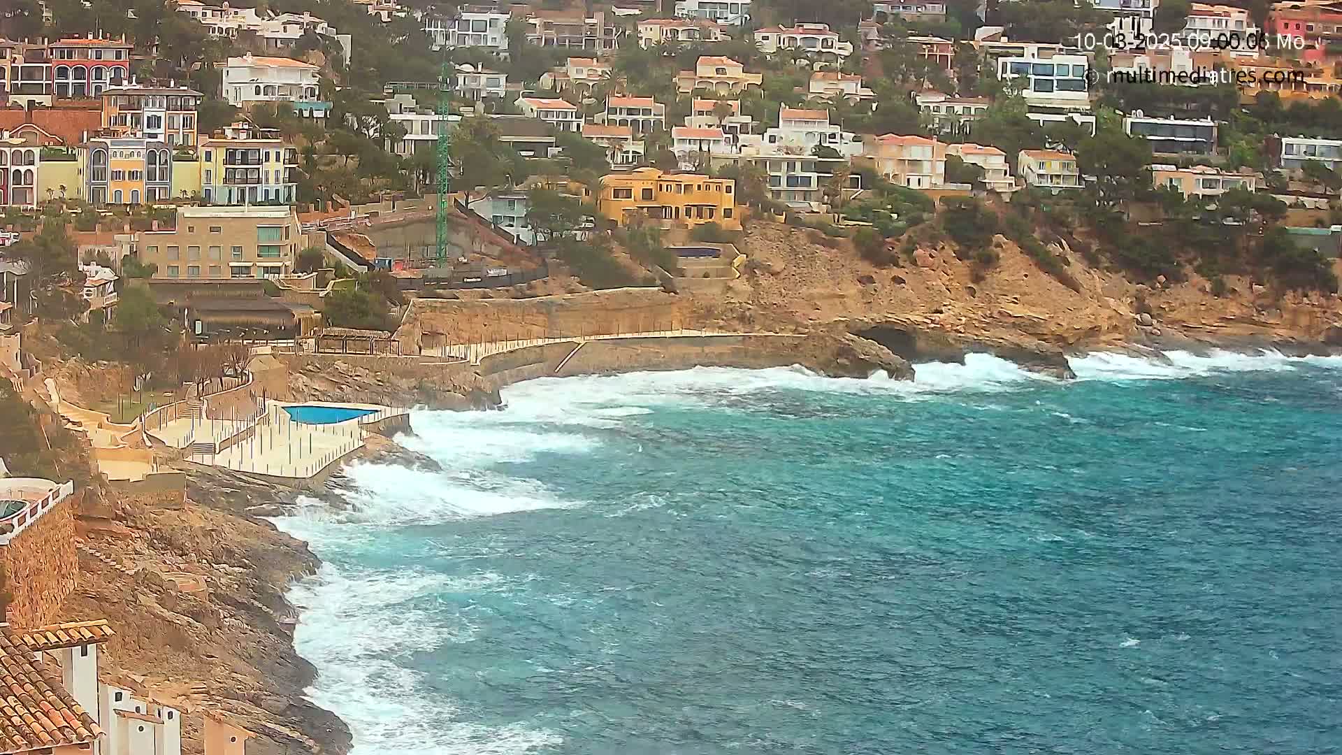 A rocky coastline with a small pool and numerous houses overlooks a choppy teal sea under overcast skies.