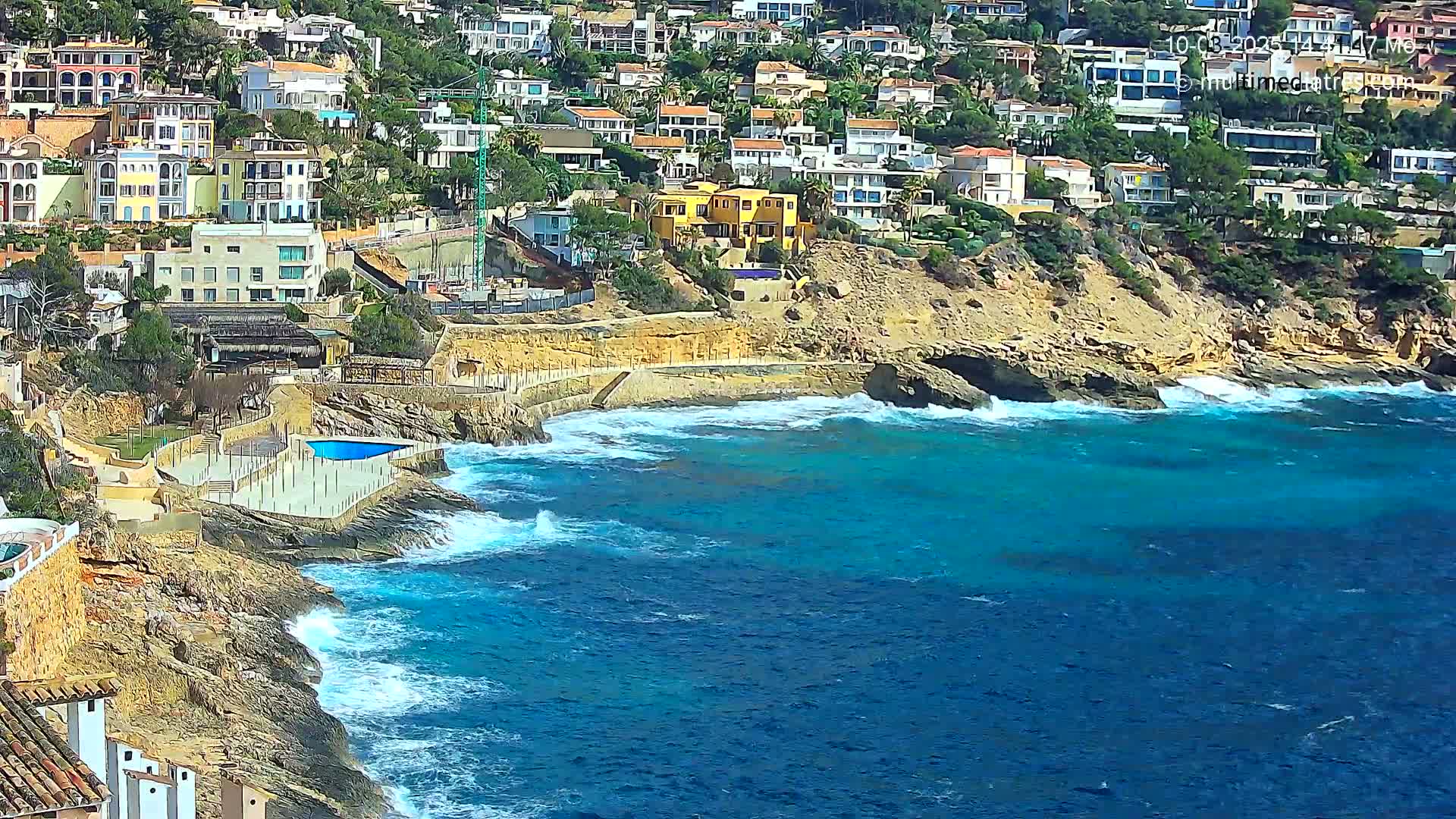 A rocky coastline with turquoise water and white-capped waves crashes against a hillside of closely packed houses.