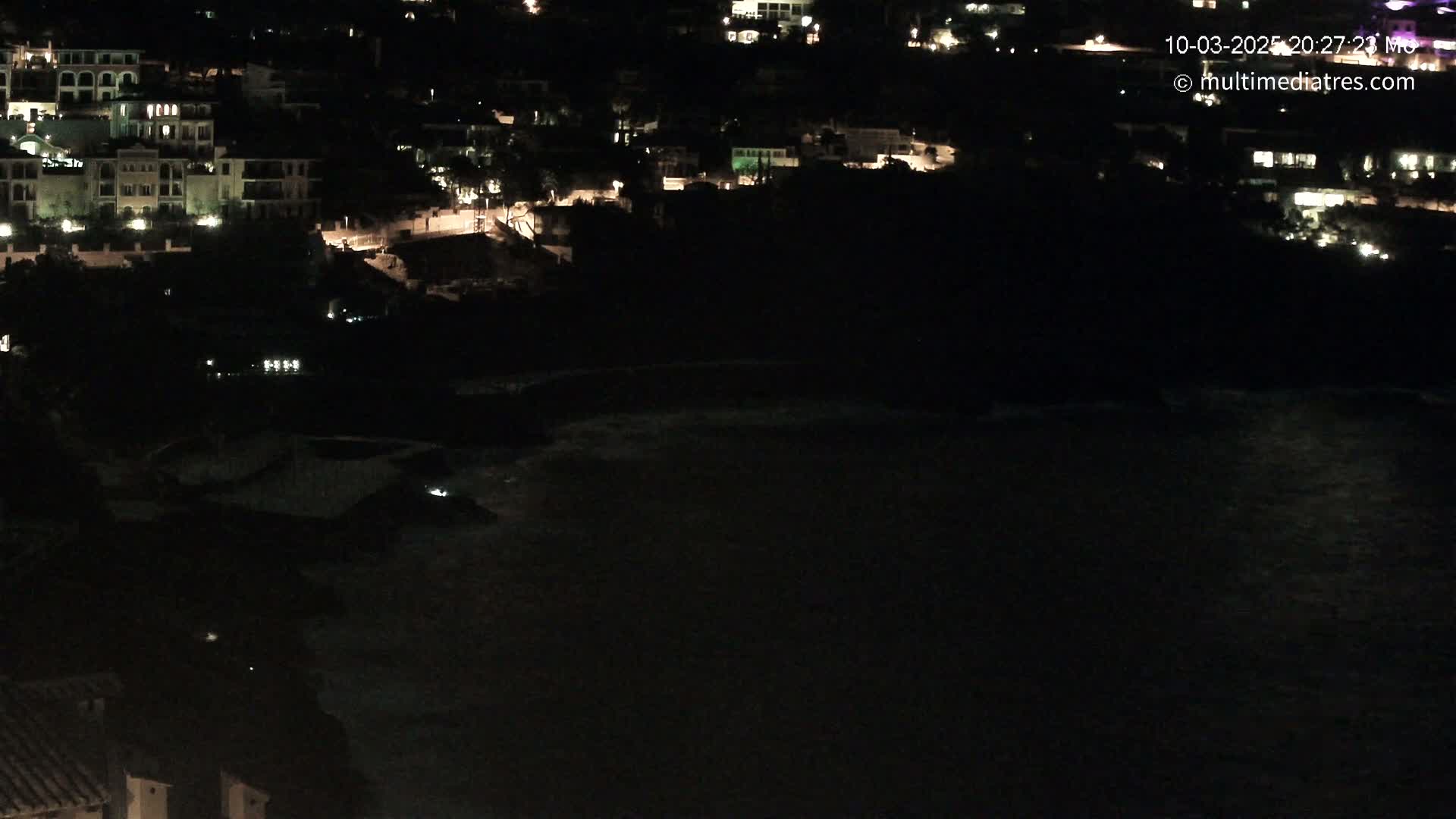 A nighttime view of a coastal town with dimly lit buildings overlooking a dark body of water with slightly visible waves.