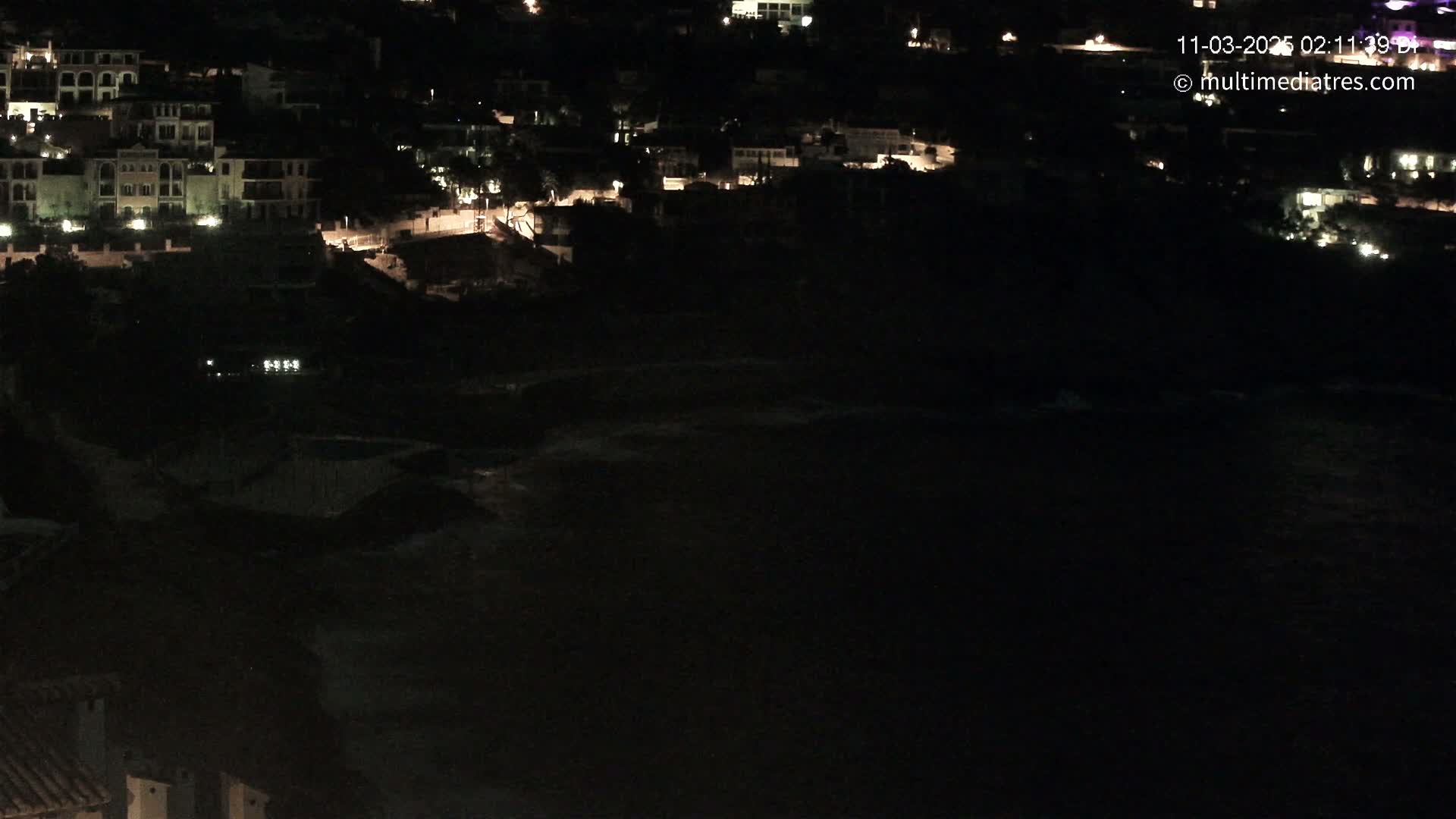 A nighttime, aerial view shows a dark coastline with faintly visible waves below a cluster of dimly lit buildings on a hillside.