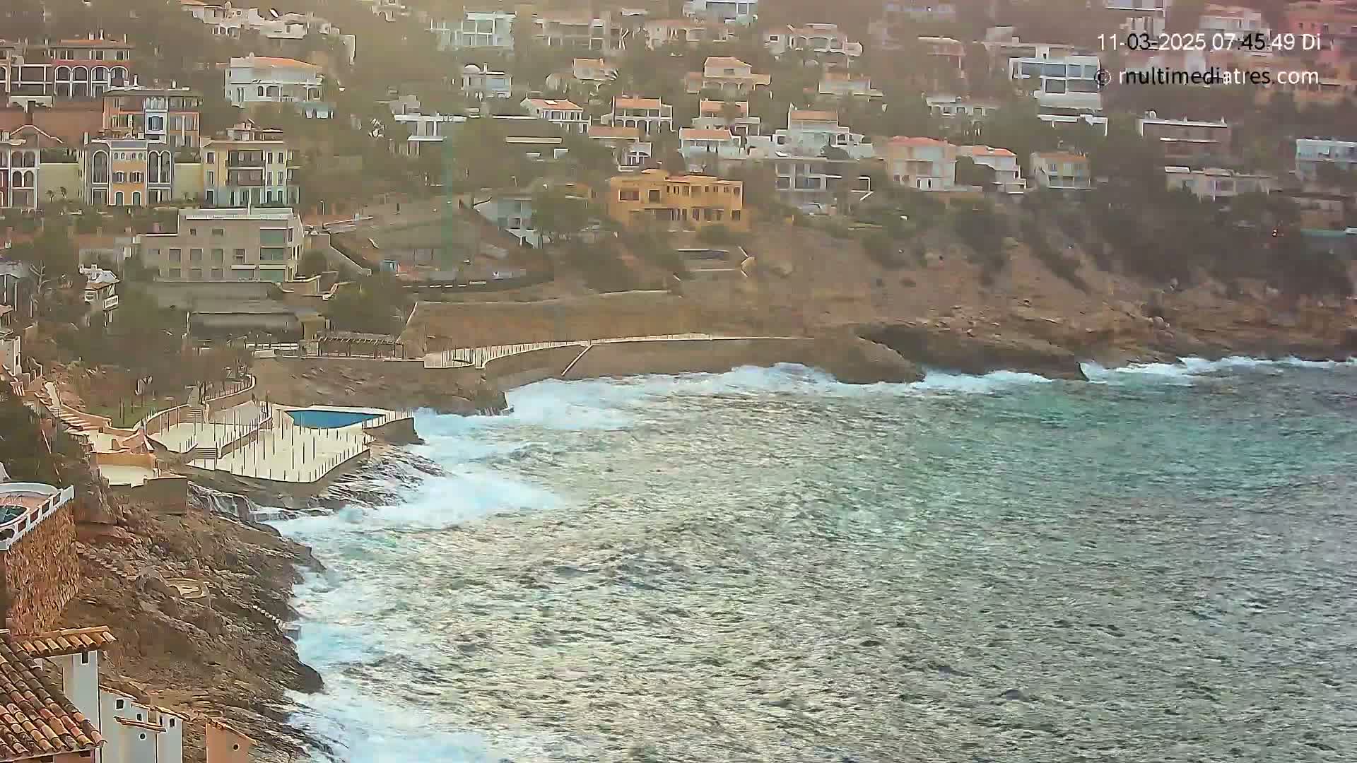 Waves crash against a rocky coastline near a cluster of buildings, overlooking a small swimming pool.
