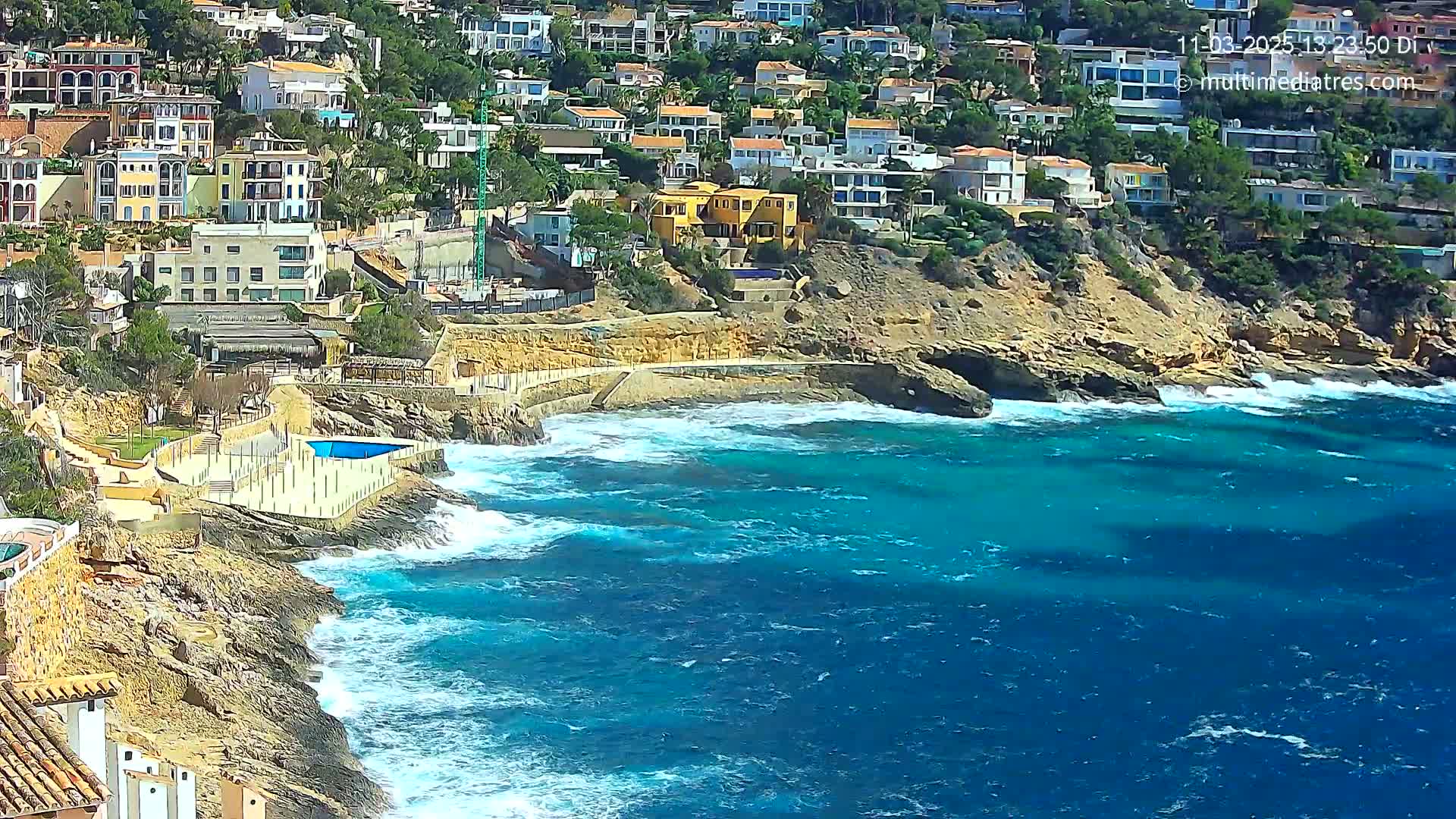 A rocky coastline with a cluster of buildings on a hillside overlooks a vibrant blue sea with moderate waves.