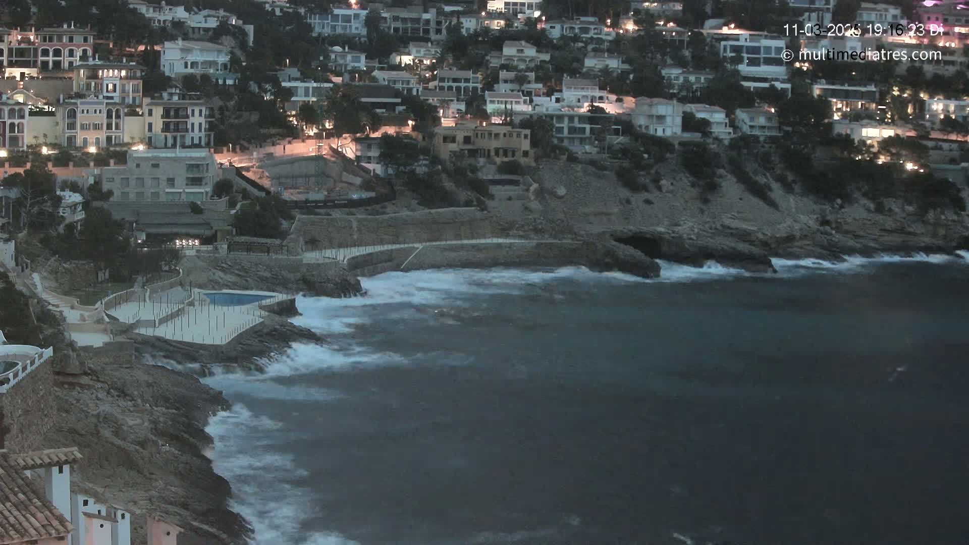 A coastal town at night with many lights on, featuring a rocky shoreline with crashing waves under a dark sky.