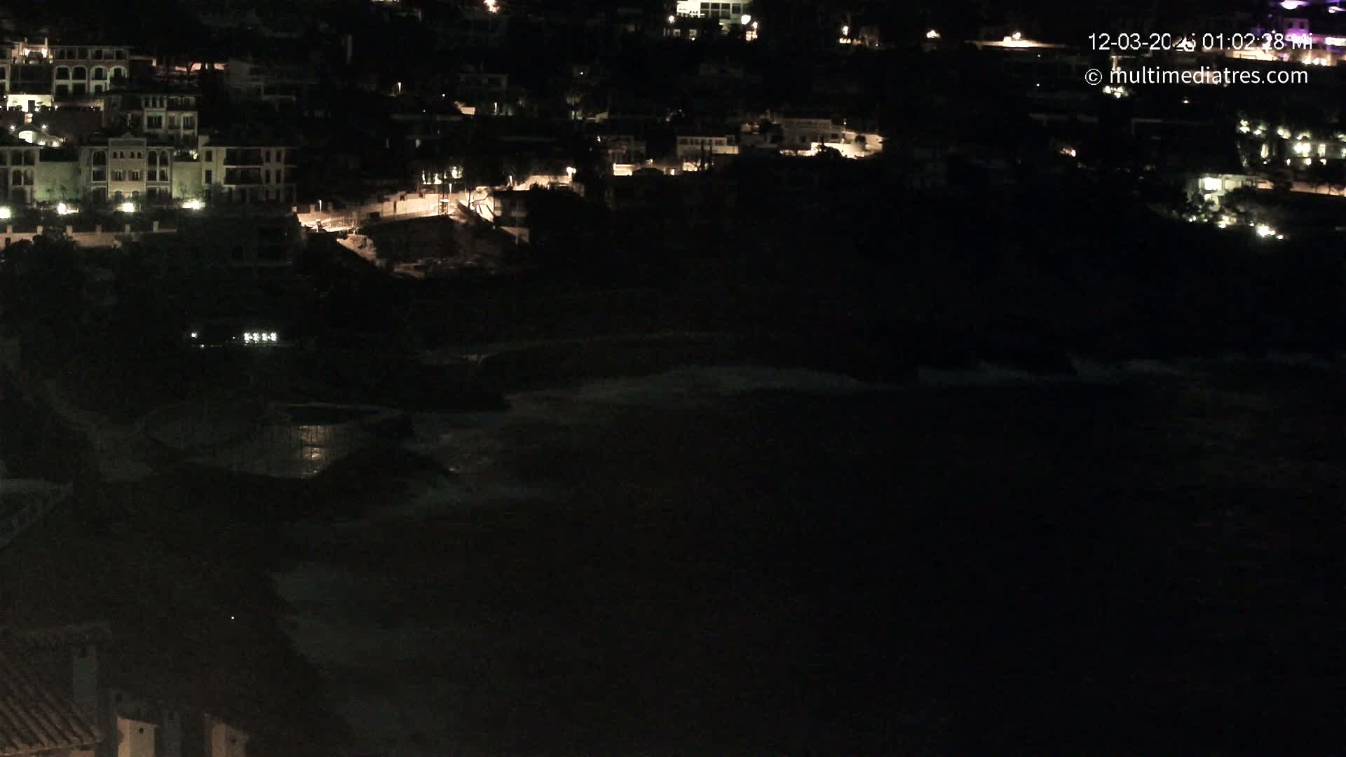 A nighttime view of a coastal town with dimly lit buildings overlooking a dark ocean with some small waves.