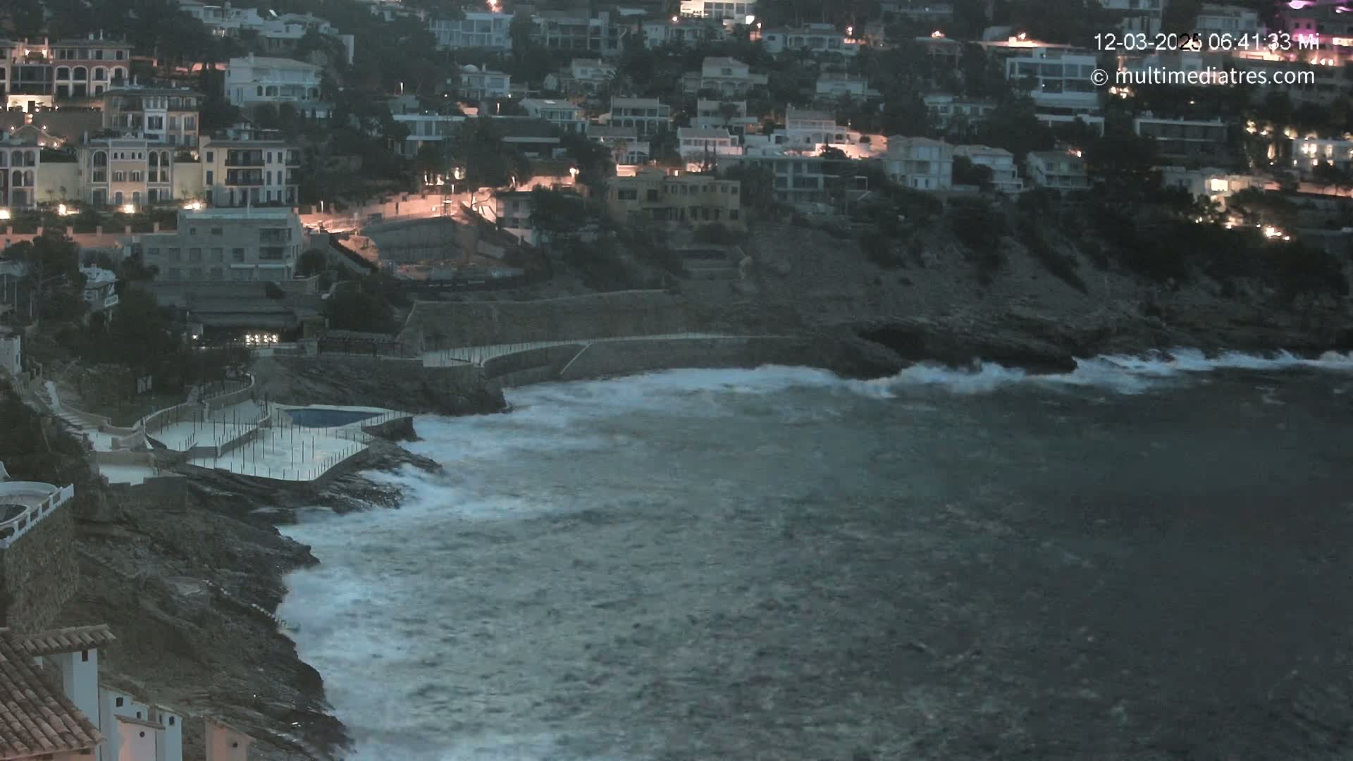 Waves crash against a rocky shoreline at night near a cluster of lit buildings.