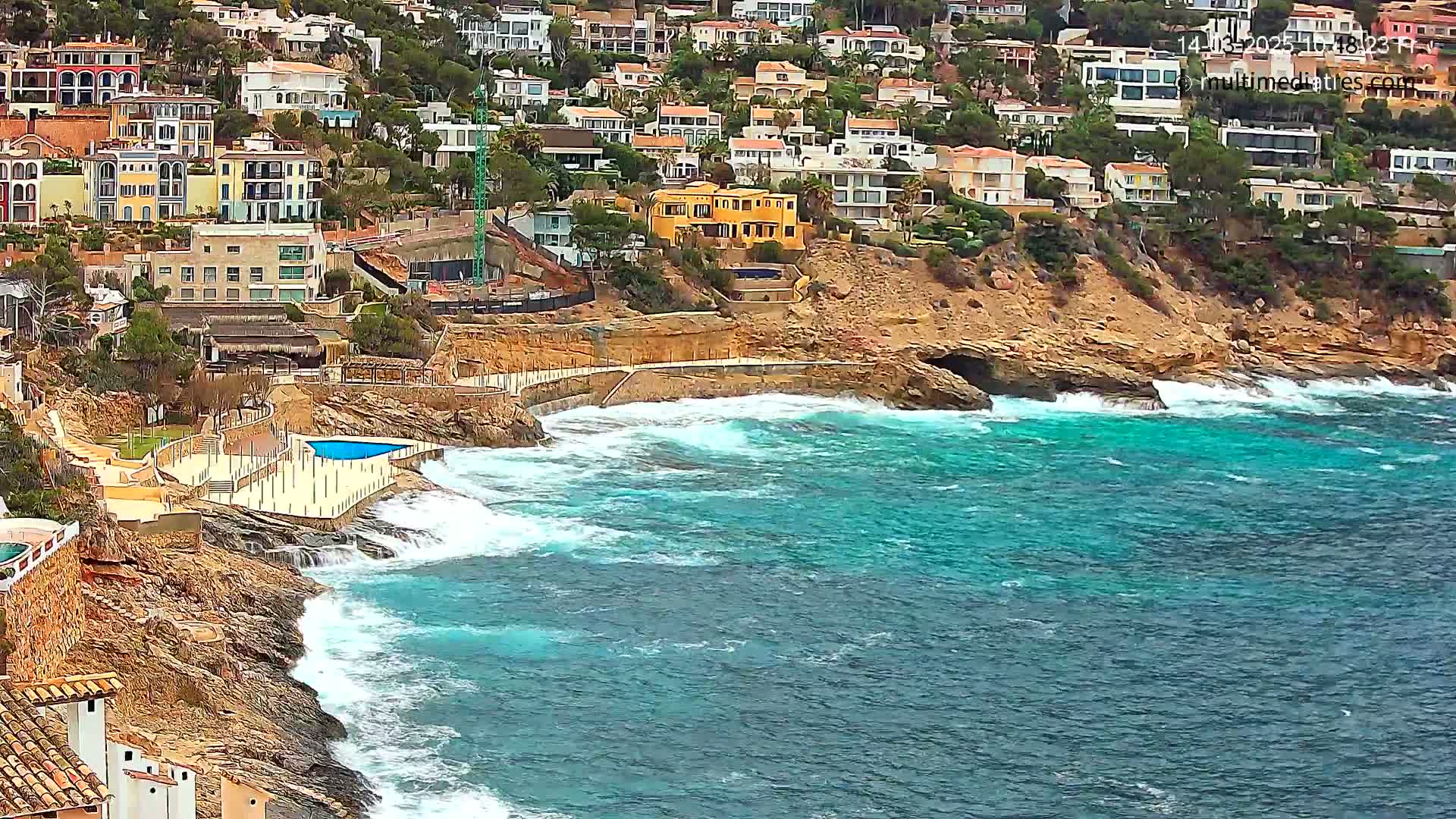 A rocky coastline with numerous houses perched on a hillside overlooks a turquoise sea with moderate waves.