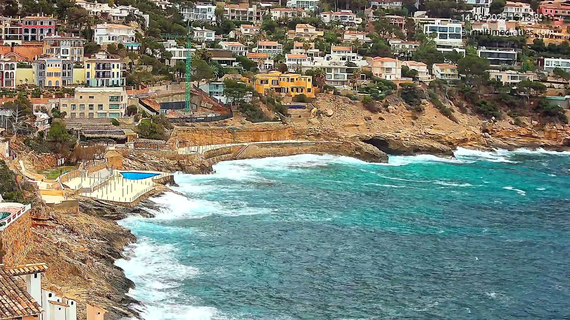 Waves crash against a rocky coastline where colorful houses cling to a hillside overlooking a turquoise sea.