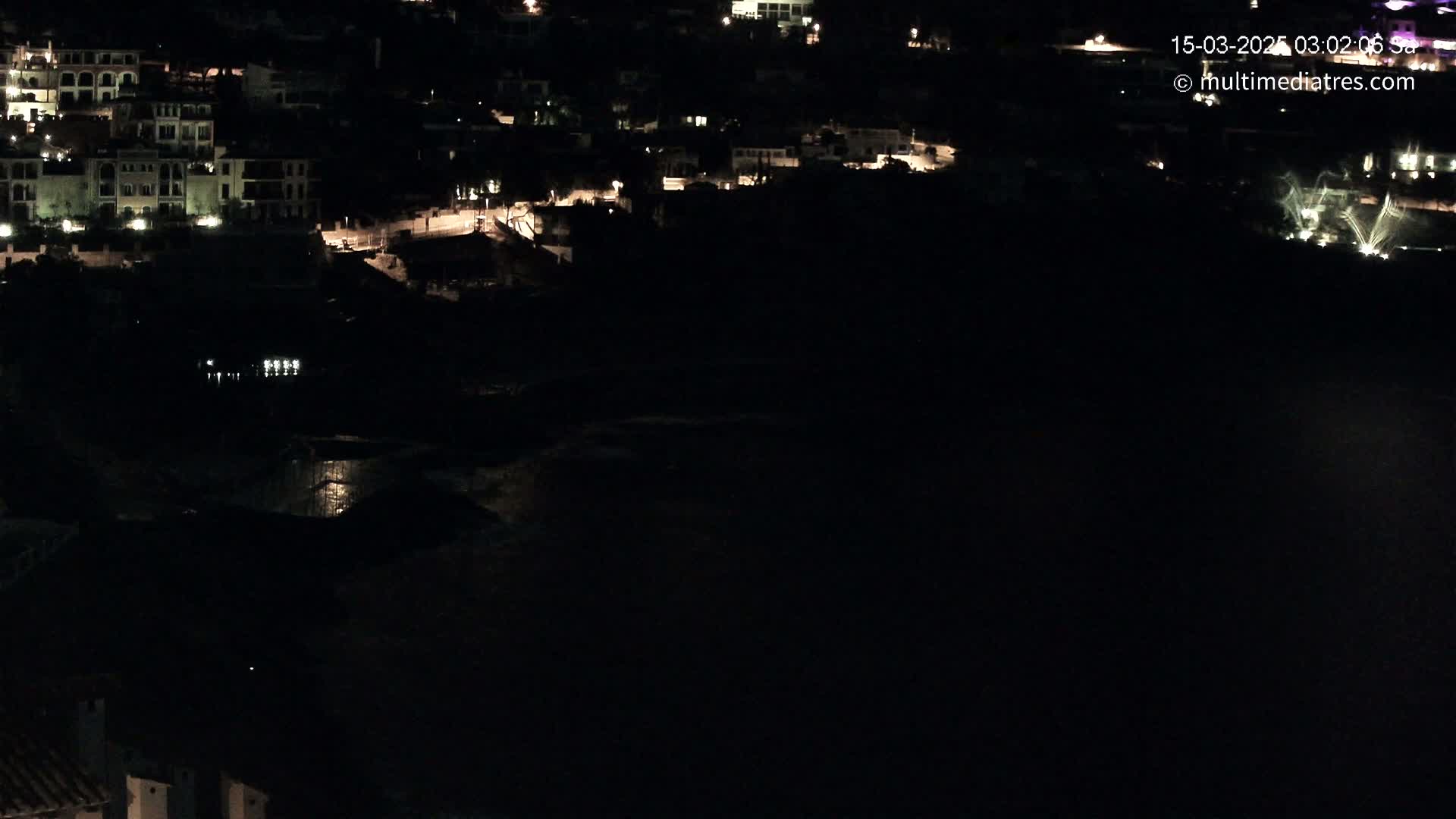 A nighttime, aerial view shows a coastal town with dimly lit buildings and a dark body of water below.