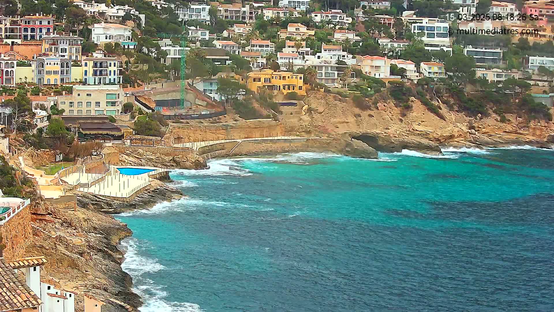 A rocky coastline with turquoise water, featuring houses built into a hillside and a small pool overlooking the ocean, under somewhat cloudy conditions.