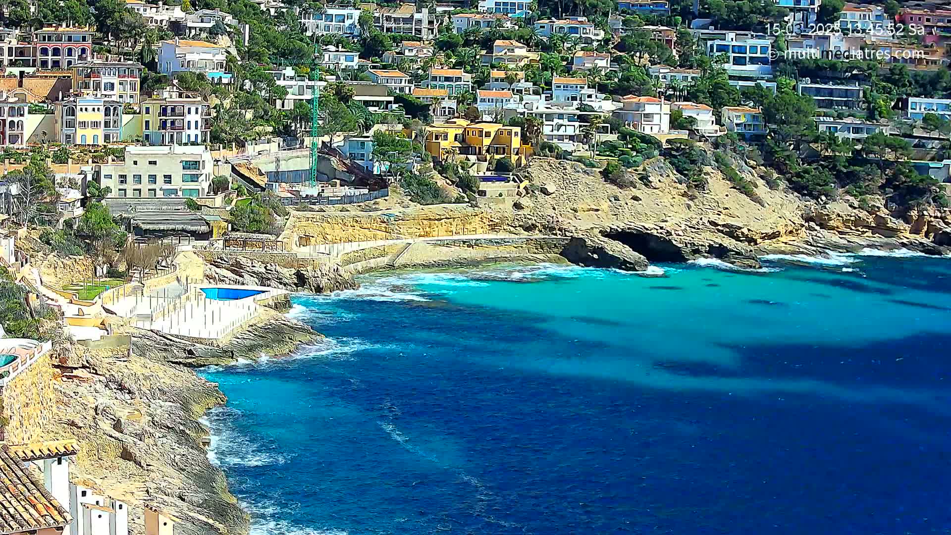 A coastal town with many houses clings to a rocky hillside overlooking a calm, bright blue sea.