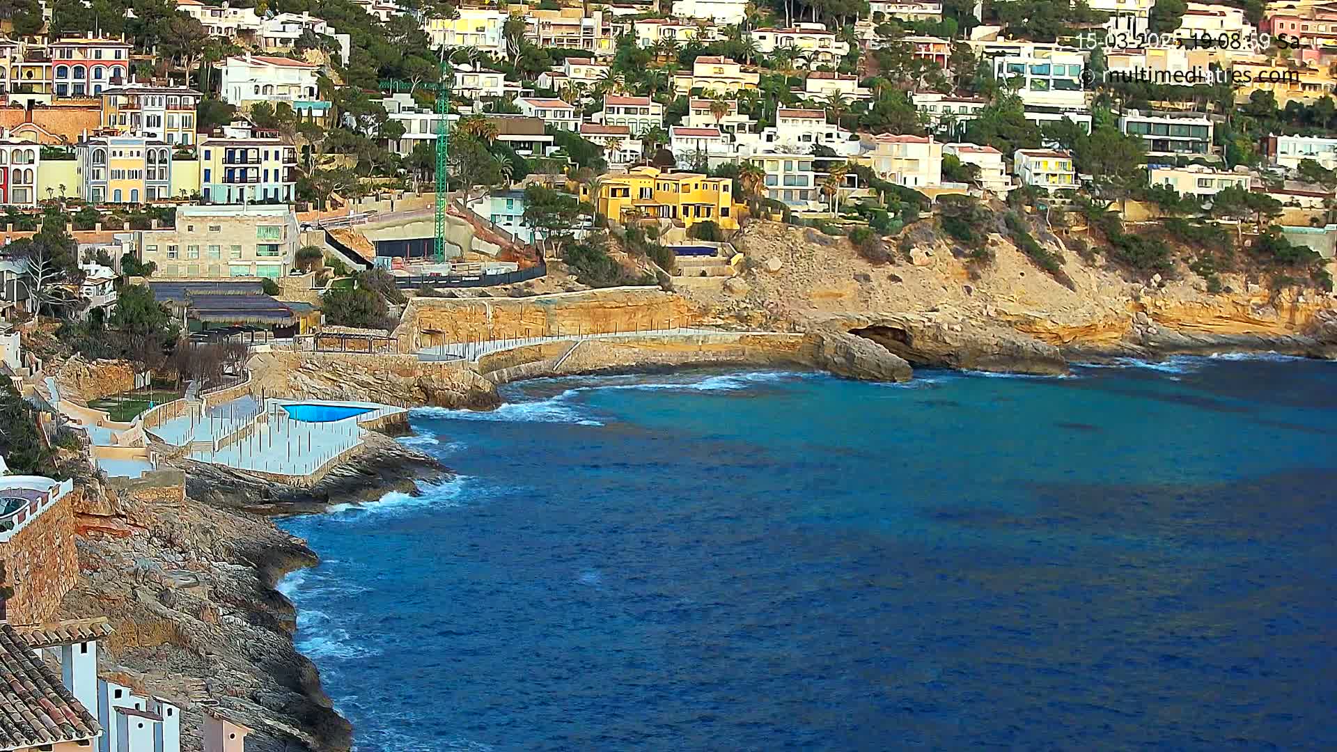 A coastal town with colorful houses built into a rocky hillside overlooks a calm, blue sea with small waves breaking on the shore.