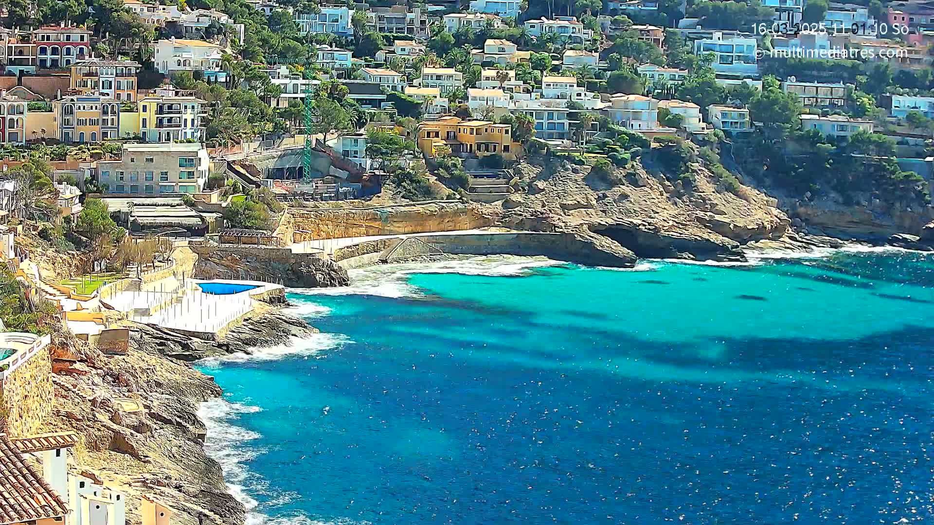 A rocky coastline with a small, turquoise cove features numerous houses built into a hillside overlooking the water on a sunny day.