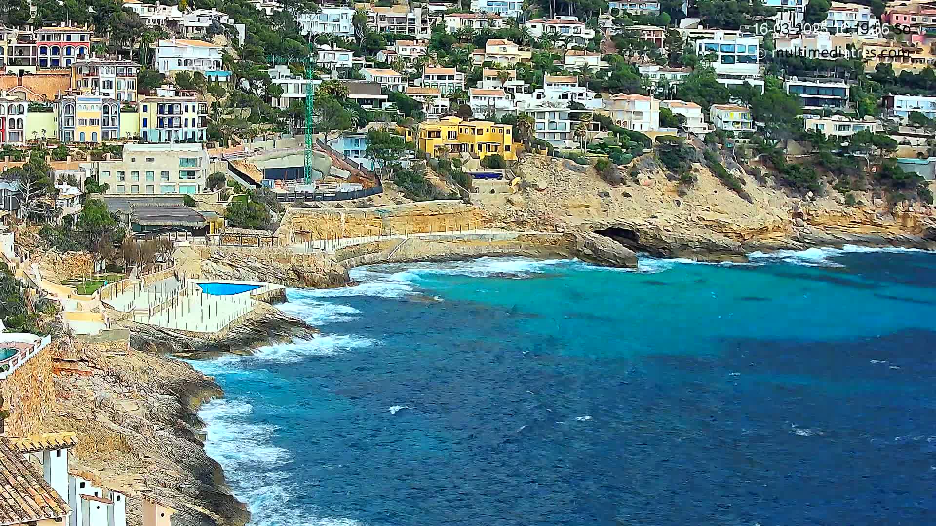A rocky coastline with a residential area overlooking a turquoise sea with small waves.
