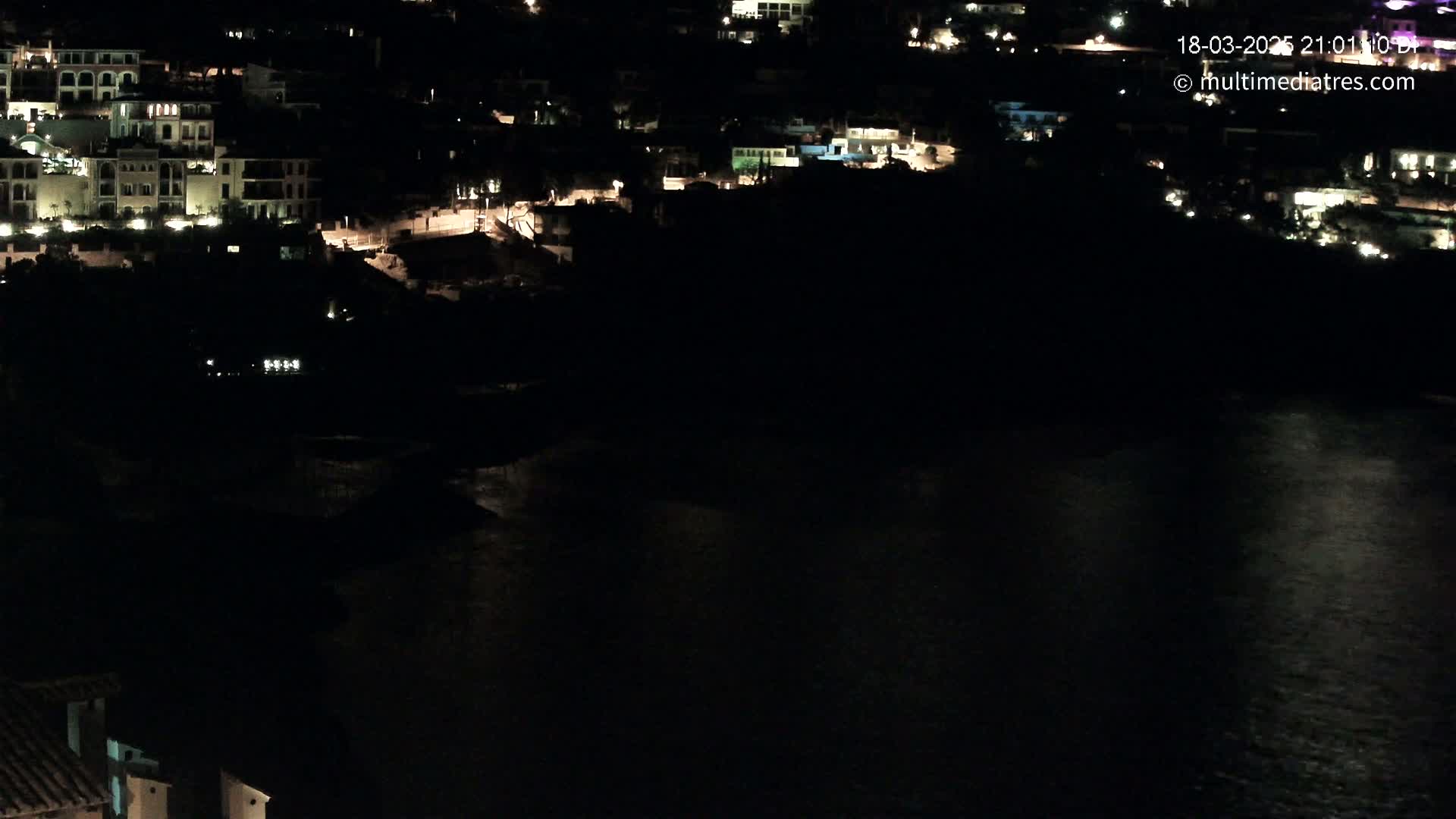 A nighttime, aerial view shows a dark body of water reflecting some lights from buildings on a hillside.