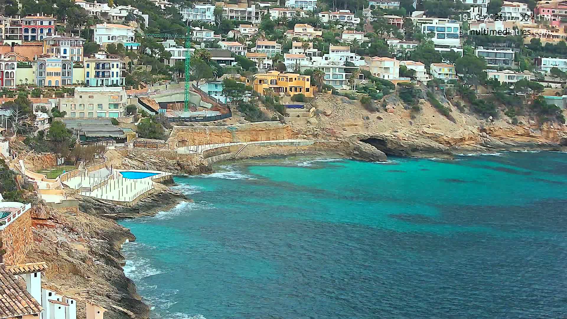 A rocky coastline with turquoise water features many houses built into a hillside, a small swimming pool, and some light wave action.