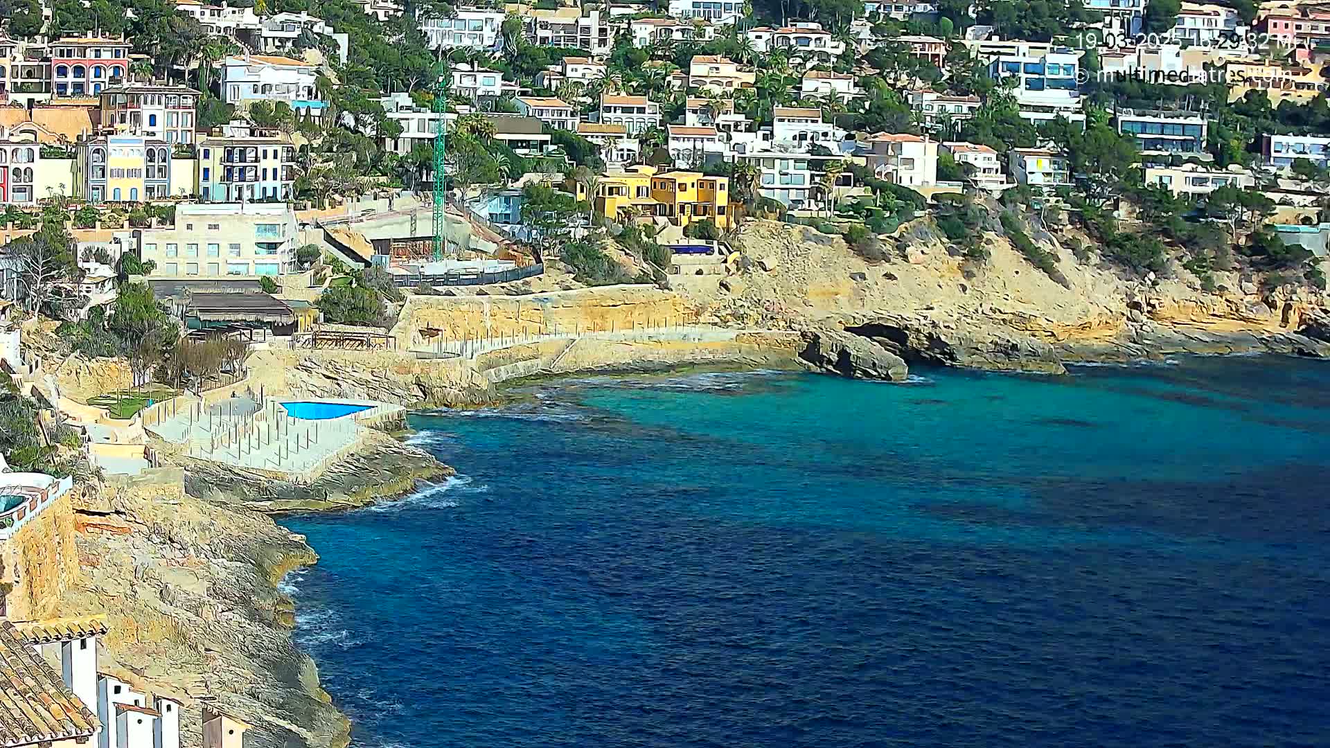 A hillside of Mediterranean-style houses overlooks a rocky coastline and a vibrant blue ocean on a sunny day.