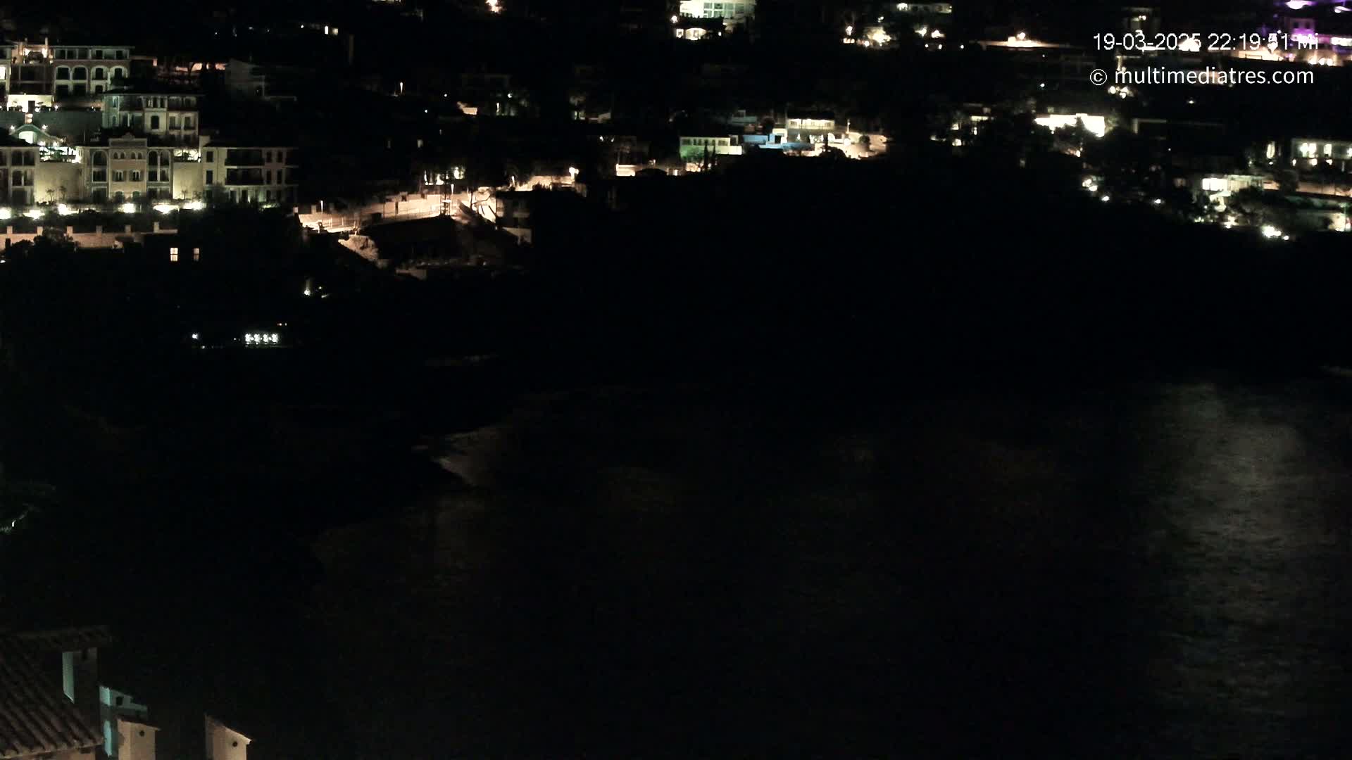 A nighttime aerial view shows a coastal town with illuminated buildings and a dark body of water reflecting some light.