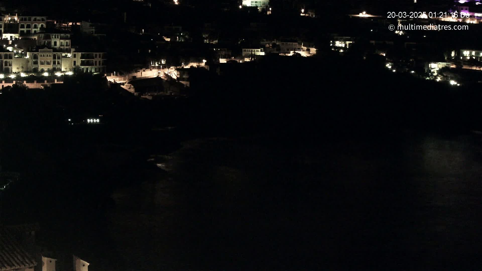 A nighttime, aerial view shows a coastal town with lit buildings near a dark body of water.