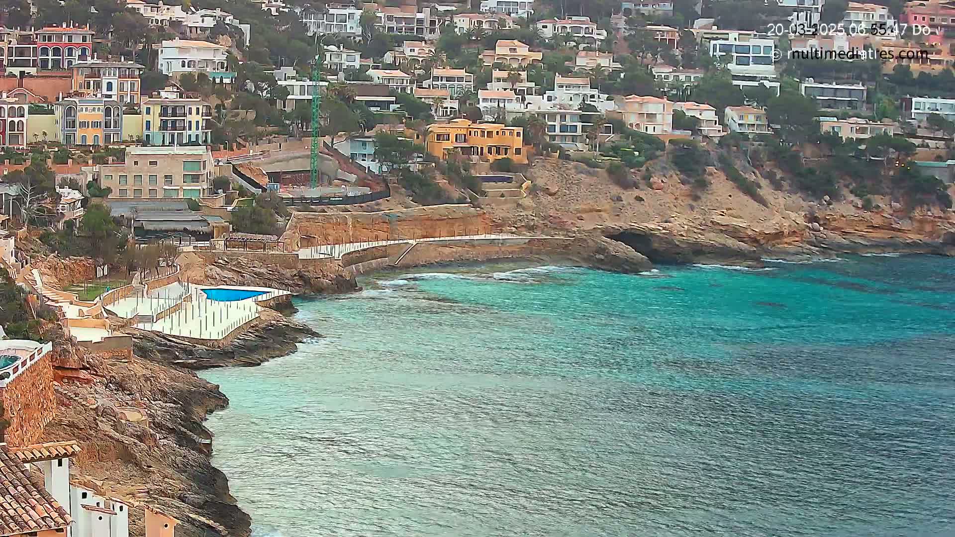 A coastal town with colorful buildings clings to a rocky hillside overlooking a calm, teal sea with a small swimming pool situated on the rocks.
