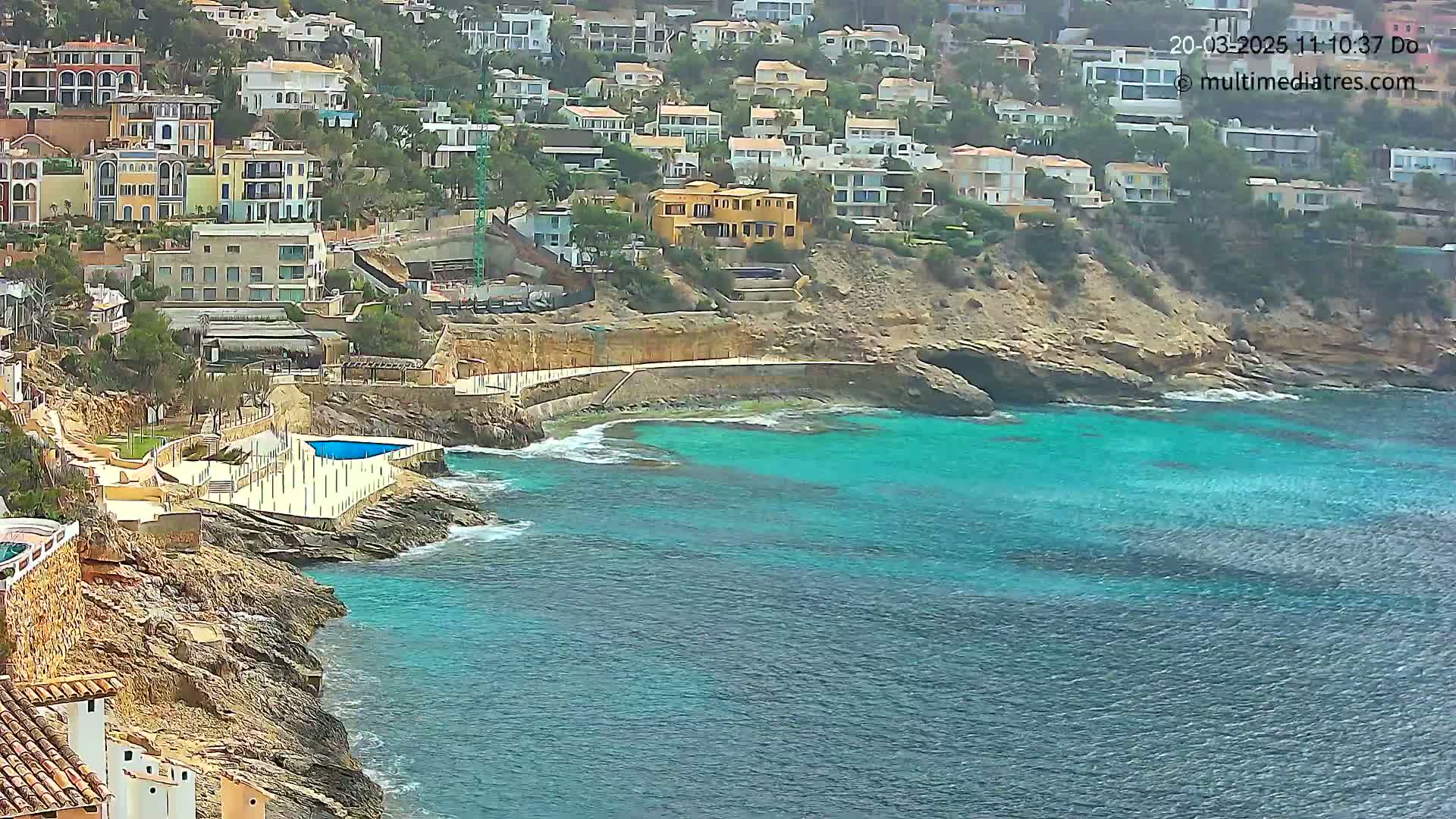 A coastal scene shows a rocky shoreline with residential buildings overlooking a teal-colored sea on an overcast day.