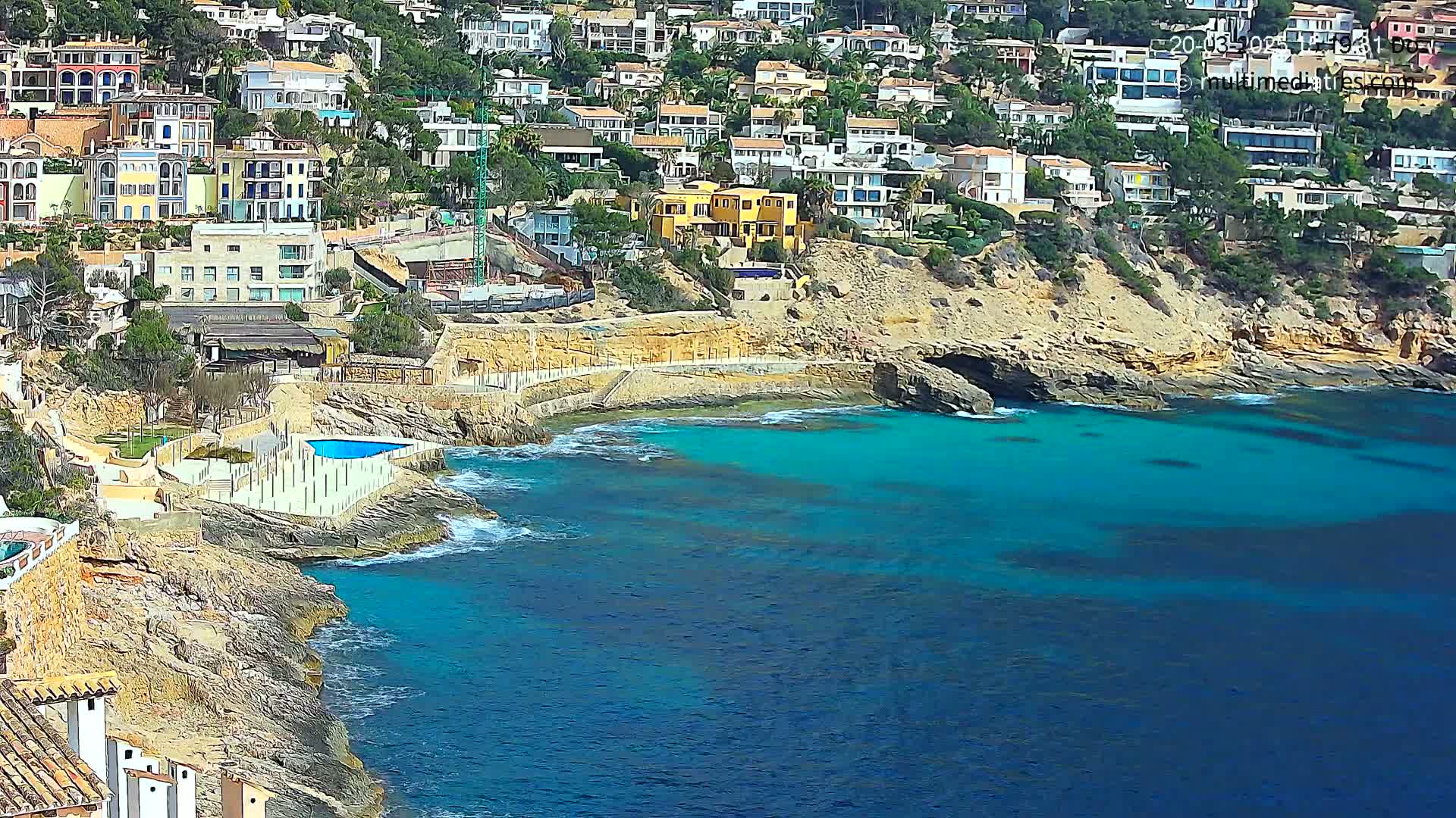 A coastal scene shows a rocky shoreline with numerous houses built into a hillside overlooking a clear, turquoise sea on a sunny day.