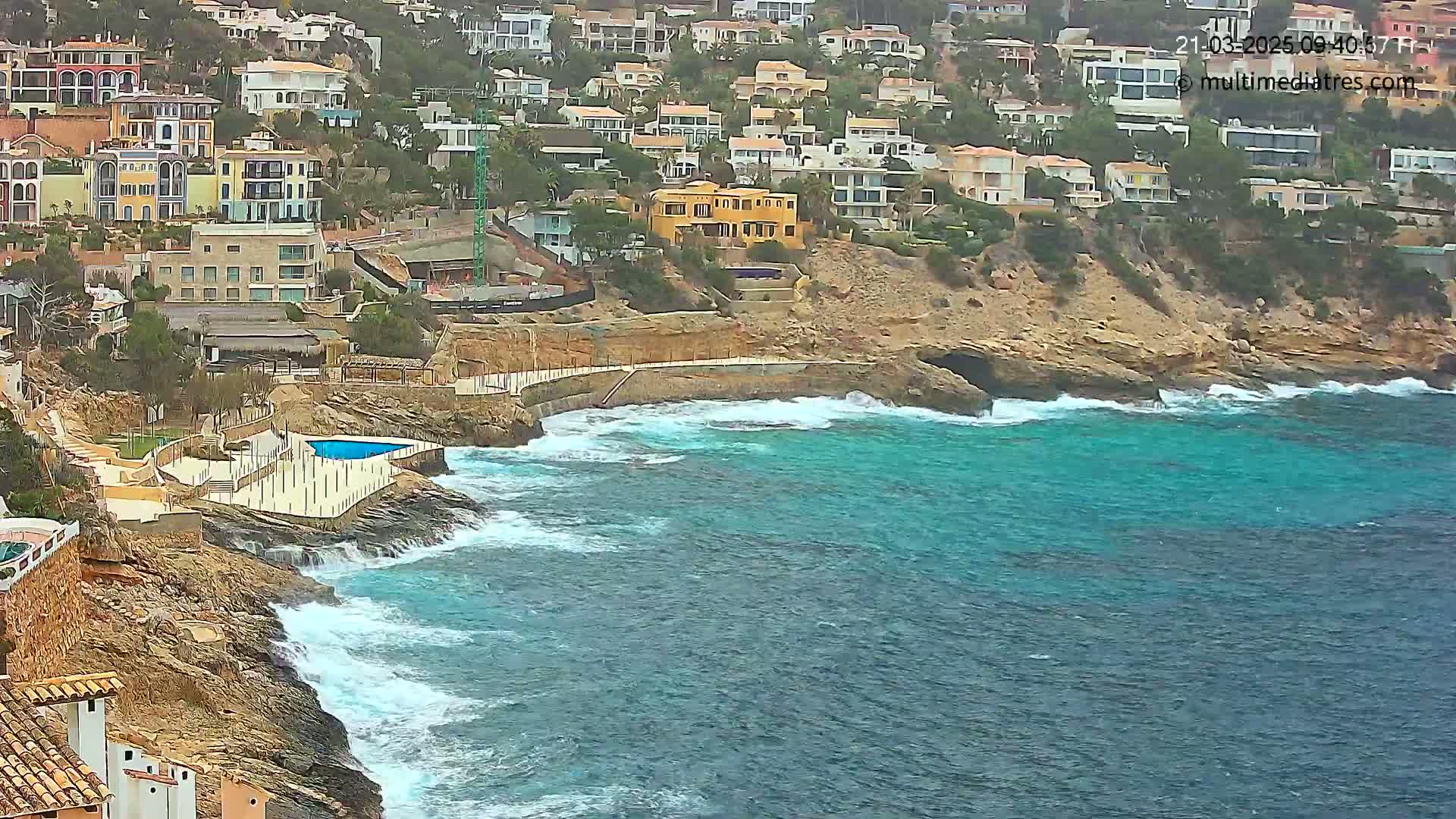 A rocky coastline with turquoise water and houses on a hillside, under somewhat overcast conditions.