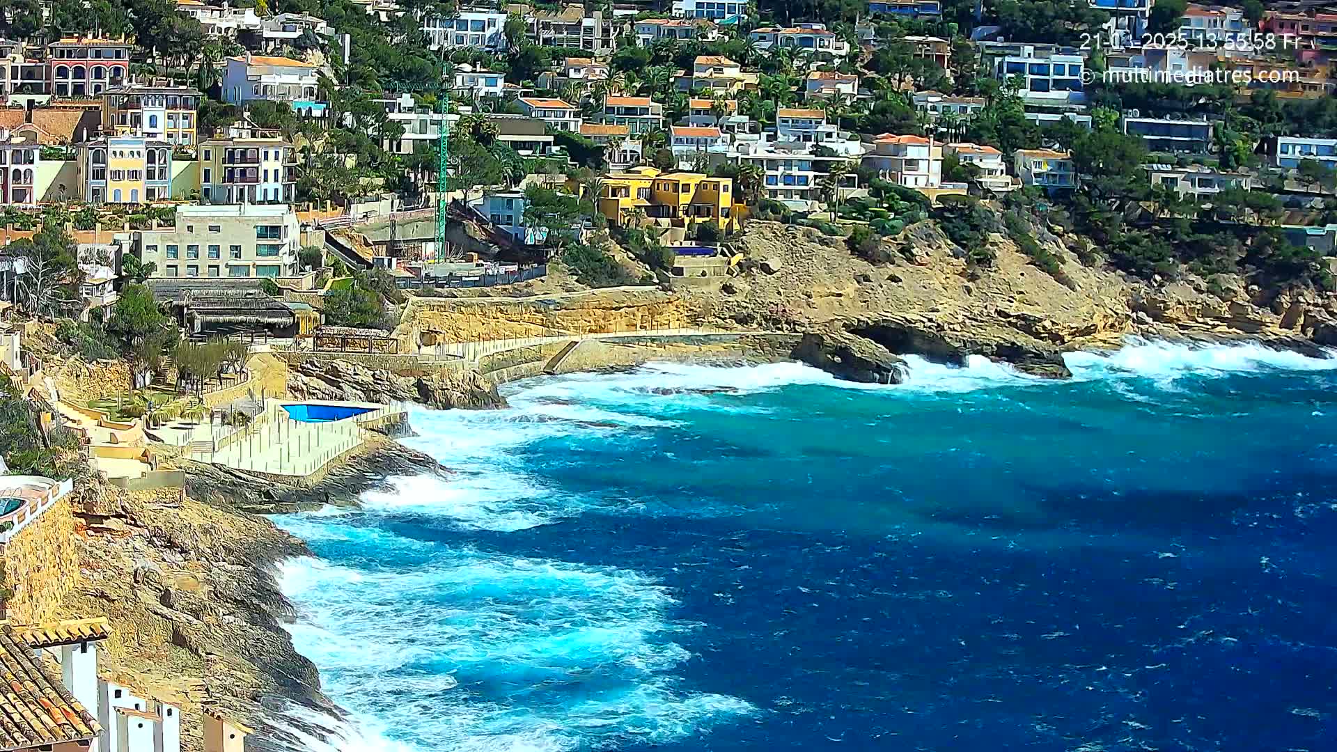 A rocky coastline with houses built on a hillside overlooks a teal sea with white-capped waves crashing against the shore on a sunny day.
