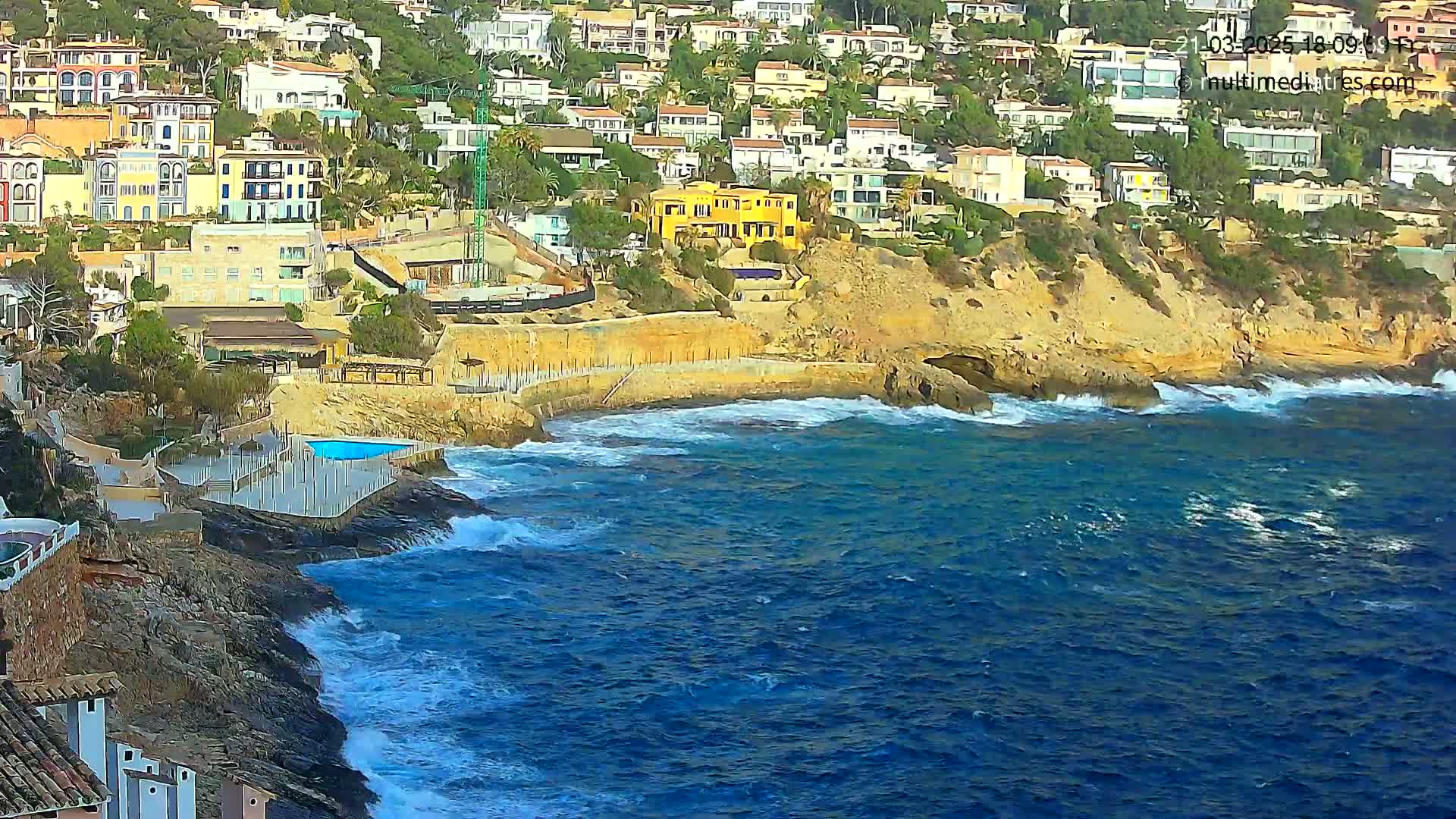 A rocky coastline with a cluster of buildings overlooks a blue sea with moderate waves.