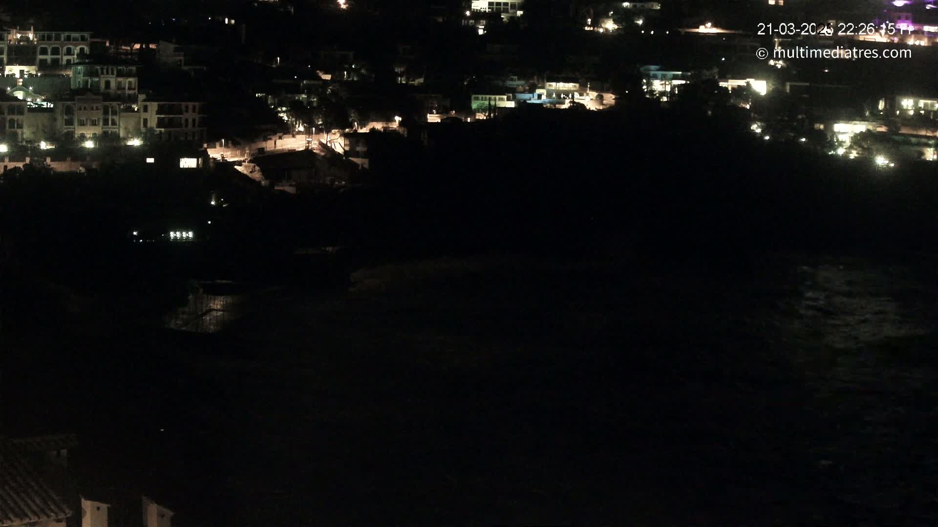 A nighttime, high-angle view shows a coastal town with scattered lights, partially obscured by darkness, and a dark body of water below.