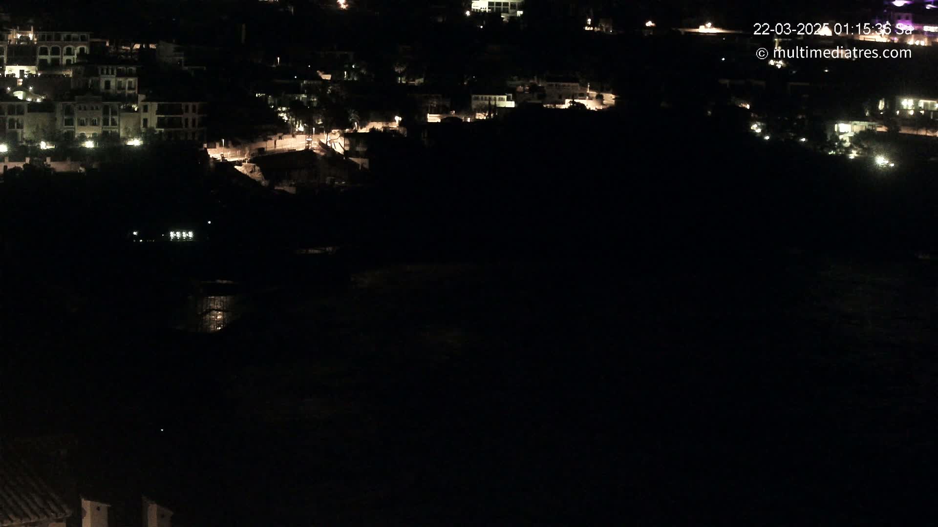 A nighttime, high-angle view shows a dark, coastal town with sparsely lit buildings and a dark body of water below.