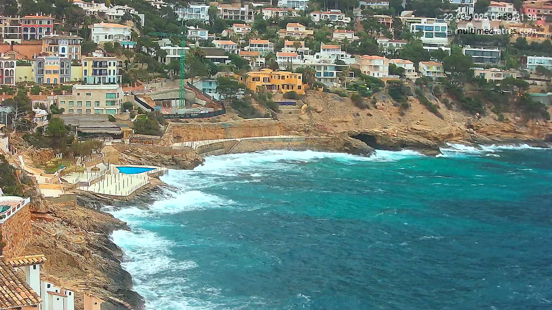 Waves crash against a rocky coastline where a cluster of buildings sits atop a cliff overlooking a turquoise sea.