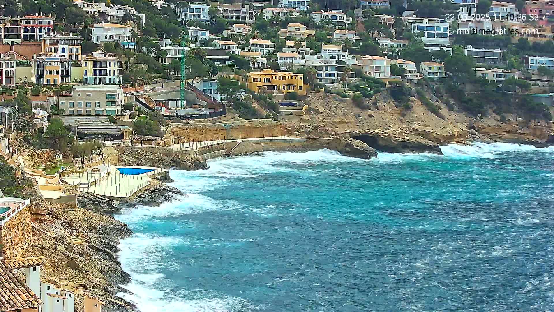 A rocky coastline with houses built into a hillside overlooks a turbulent turquoise sea with crashing waves.