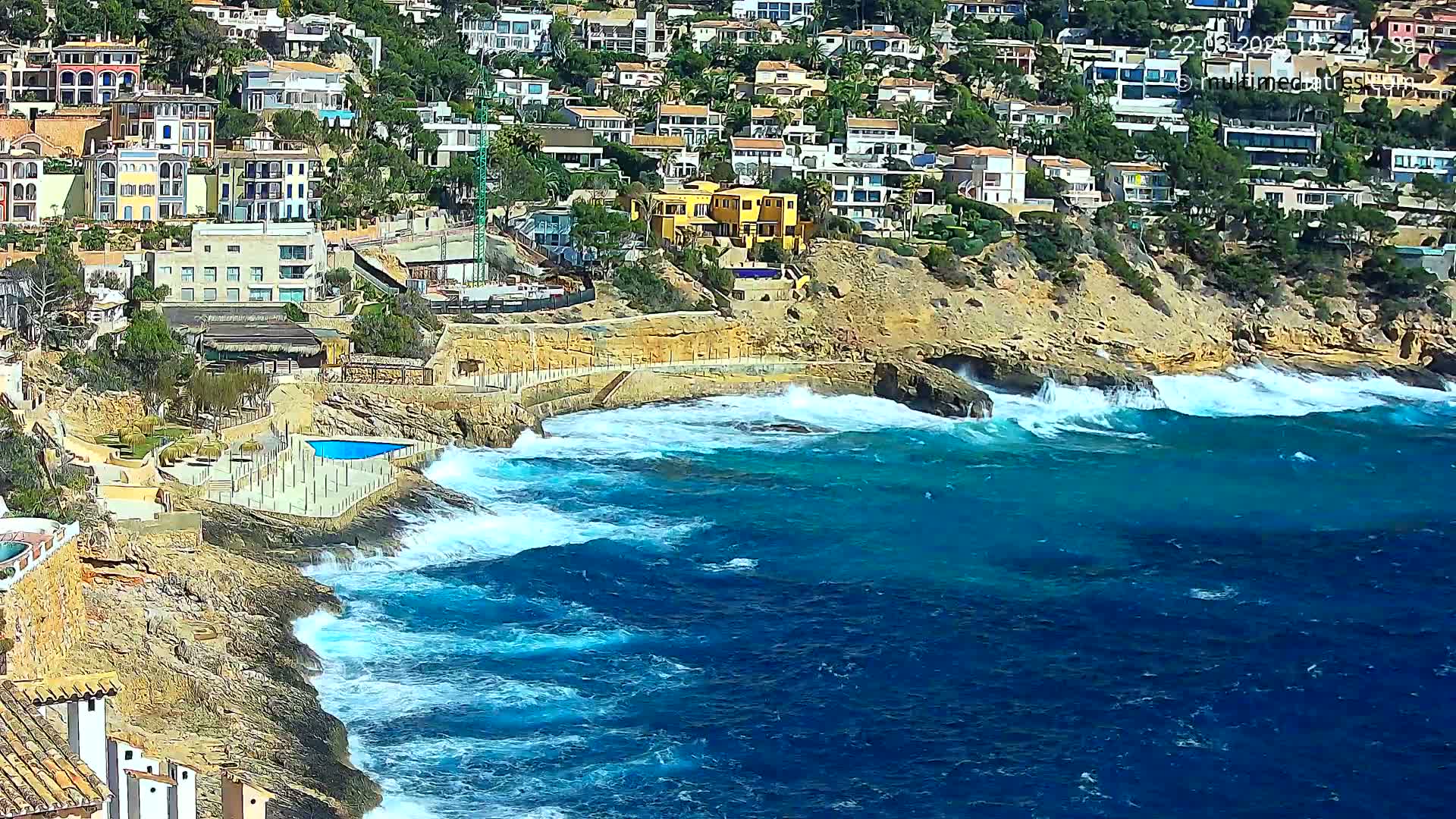 A rocky coastline with houses built into the hillside overlooks a turbulent blue sea with crashing waves.