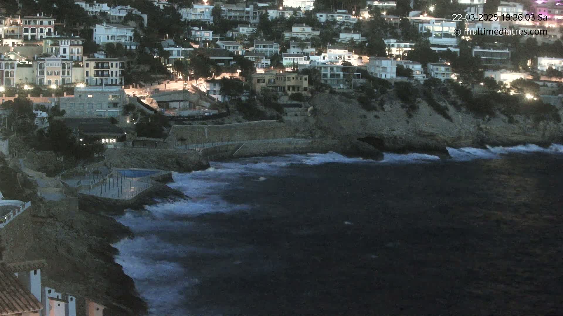 A rocky coast at night with moderately rough waves crashing against a seawall, next to a cluster of lit buildings.