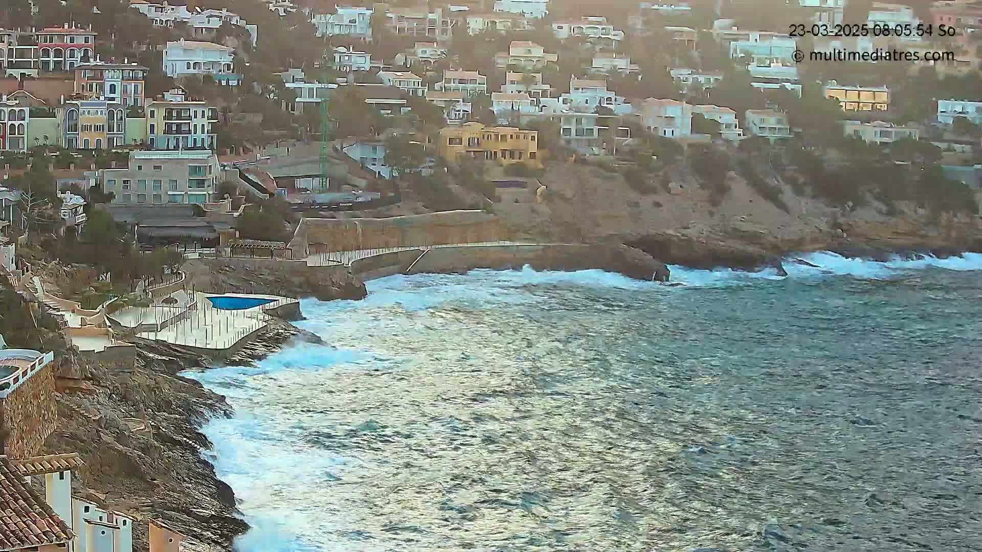 Waves crash against a rocky coastline where houses are built into the hillside.