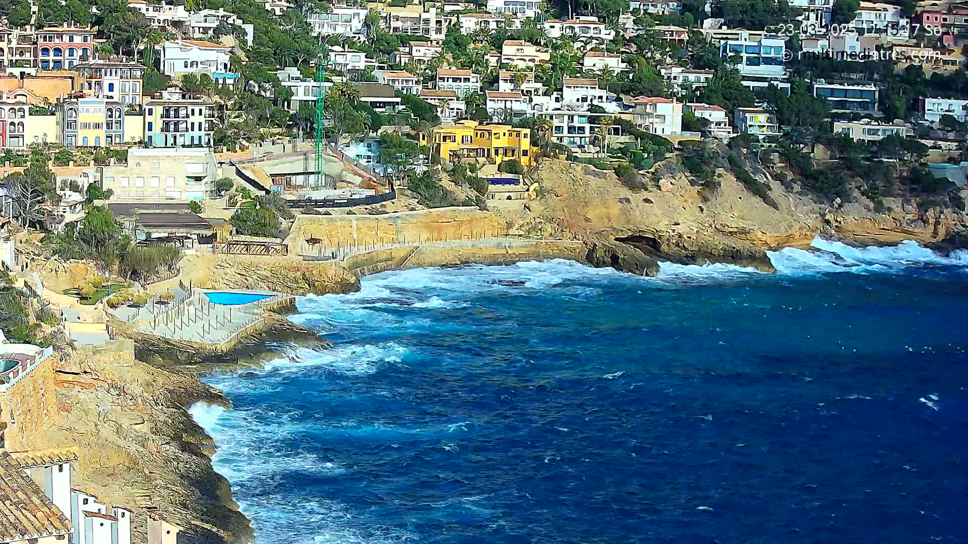 A rocky coastline with houses built into the hillside meets a blue ocean with moderate waves.