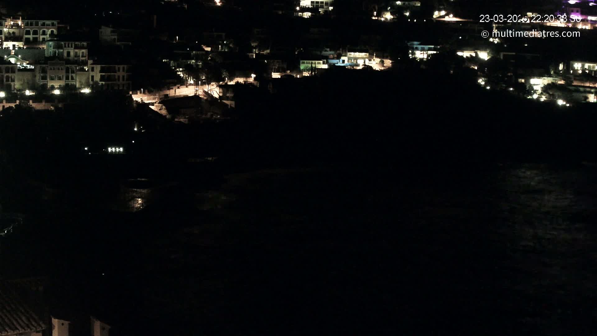 A nighttime, coastal town with sparsely lit buildings is seen from an elevated vantage point overlooking dark water.