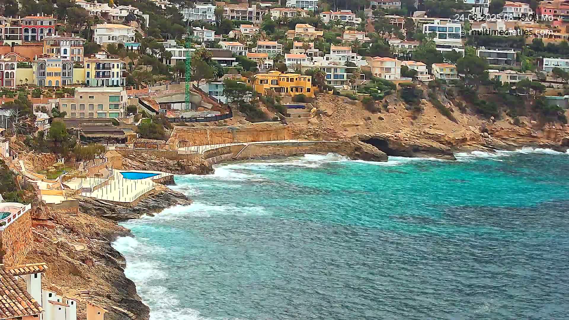 A rocky coastline with turquoise water and buildings on a hillside, featuring small waves breaking against the shore.