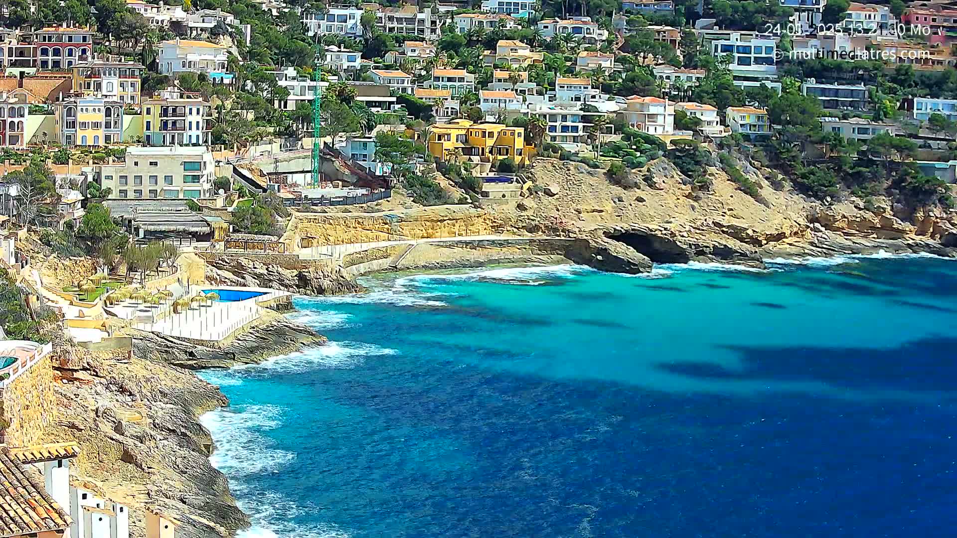 A rocky coastline with colorful houses on a hillside overlooks a clear, turquoise ocean with gentle waves.