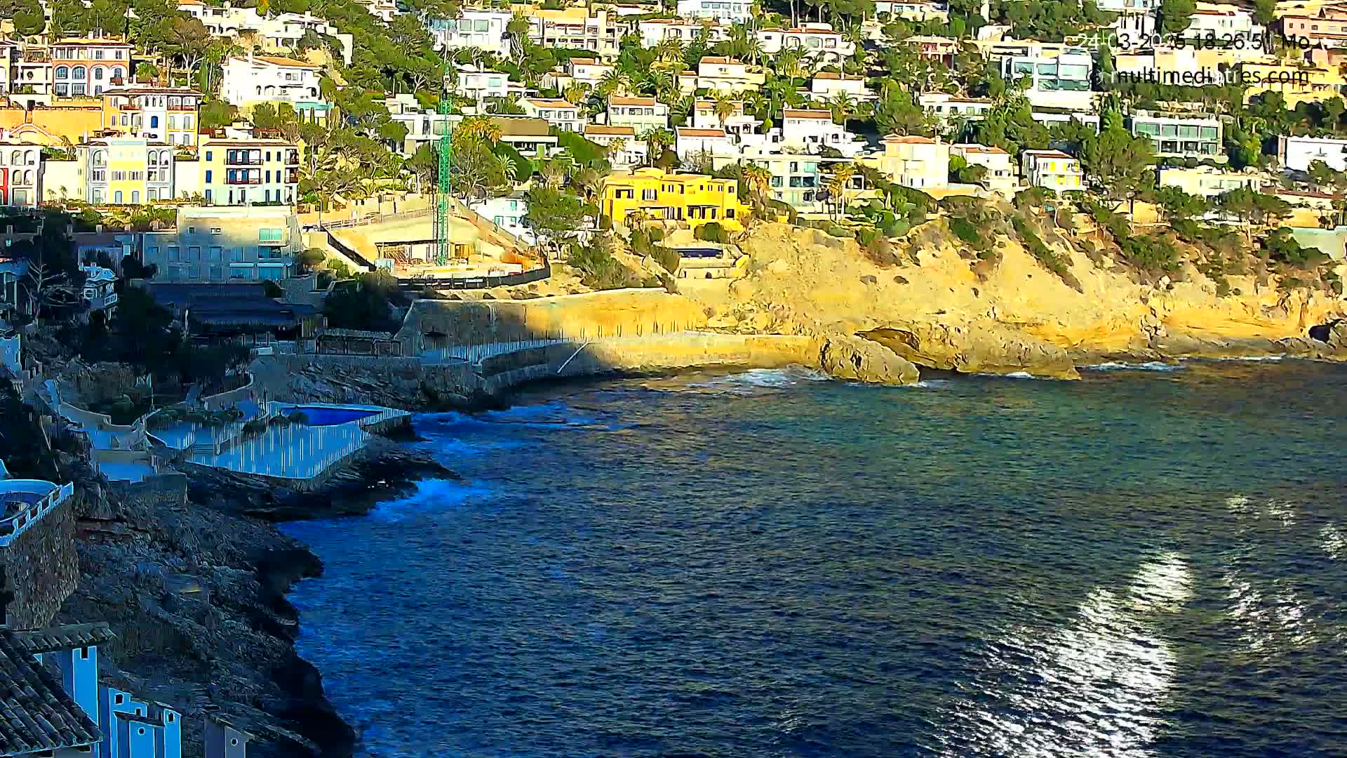 A coastal scene shows a cluster of buildings on a cliff overlooking a dark blue sea with small waves and bright reflections.