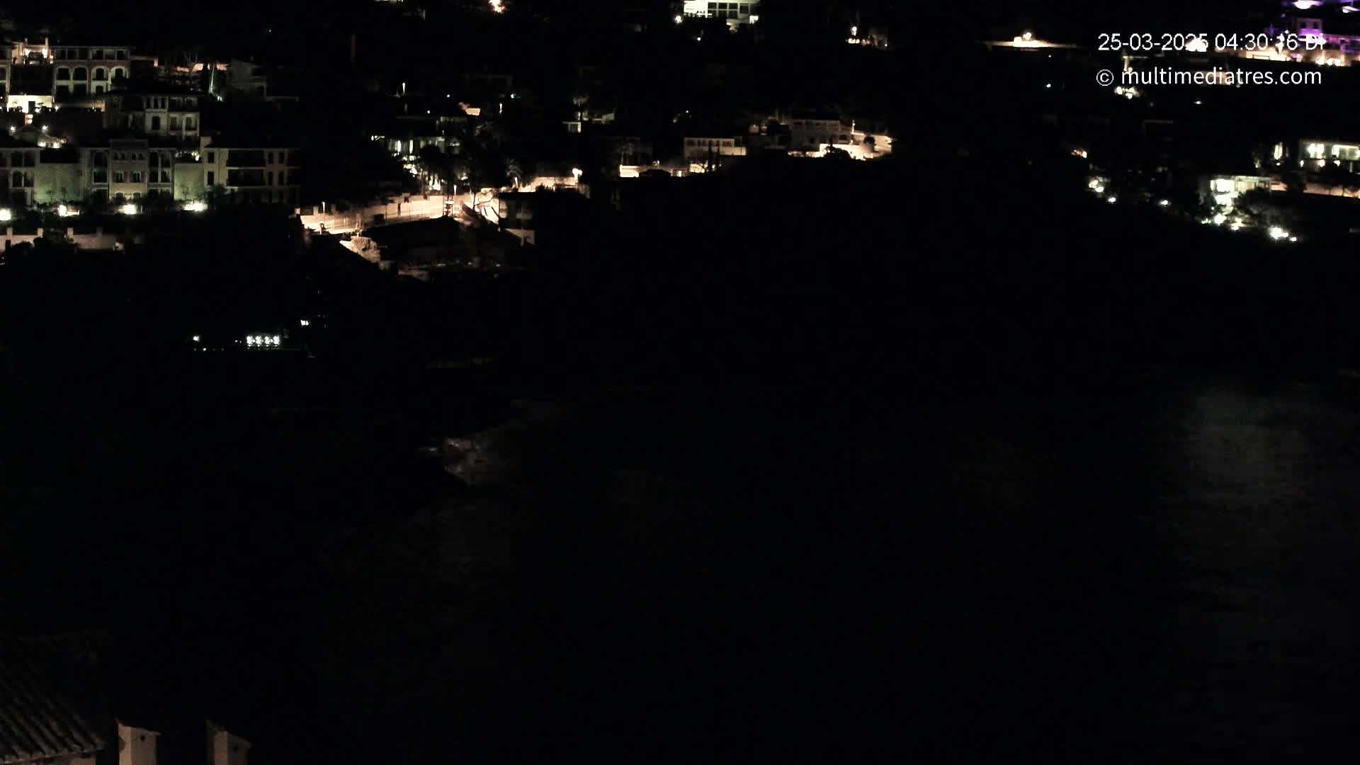 A nighttime, aerial view shows a dark body of water below a cluster of dimly lit buildings on a hillside.