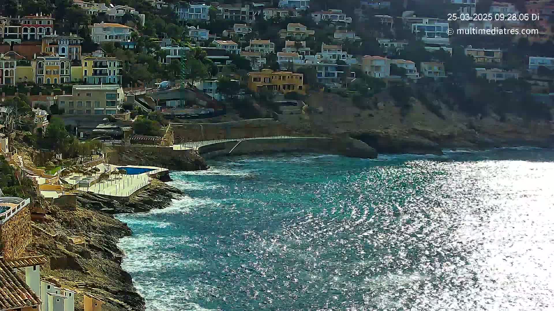 A coastal town with many houses built on a cliff overlooking a sparkling blue sea with moderate waves.