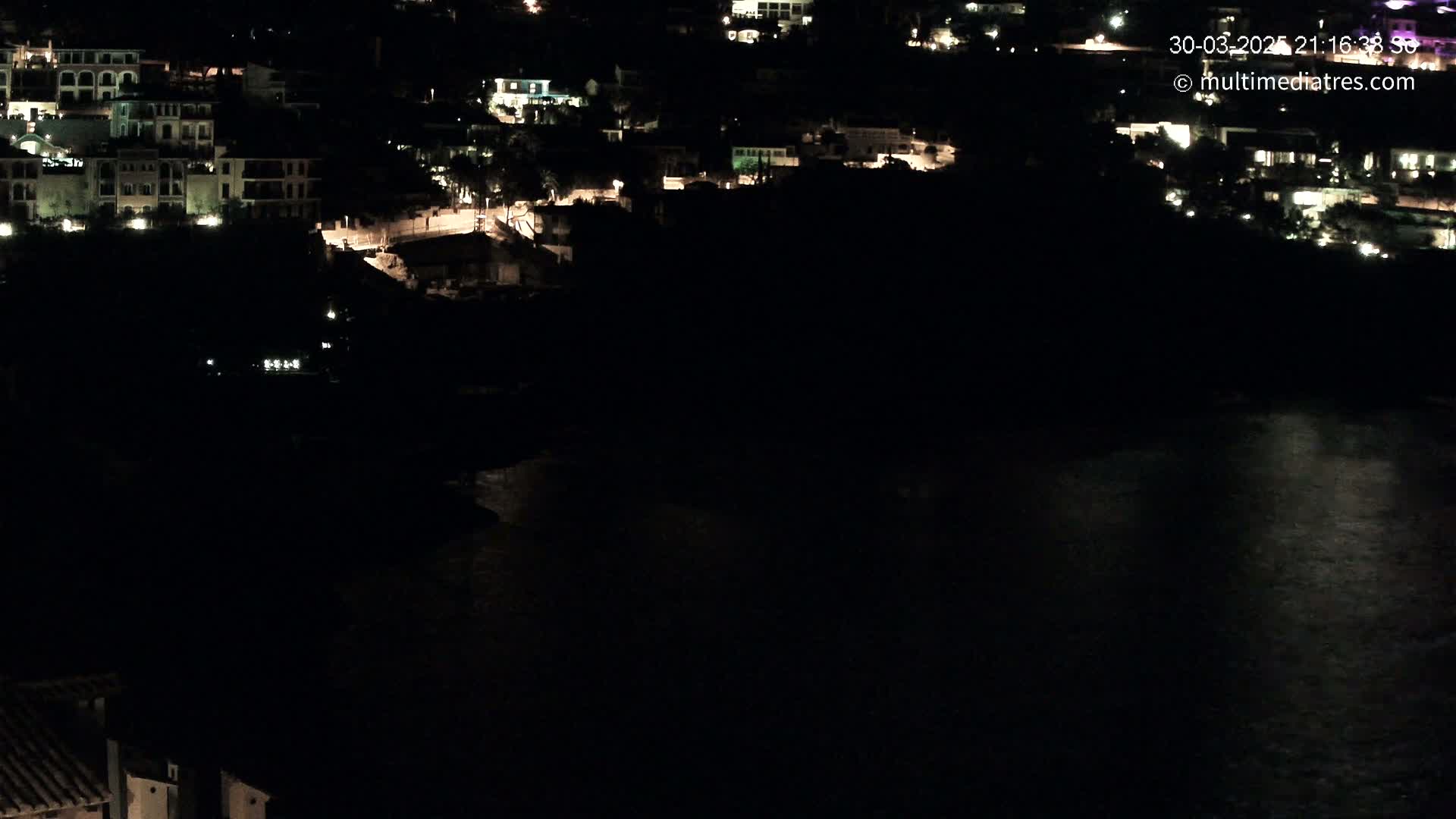 A nighttime view of a coastal town with dimly lit buildings and a dark body of water below.