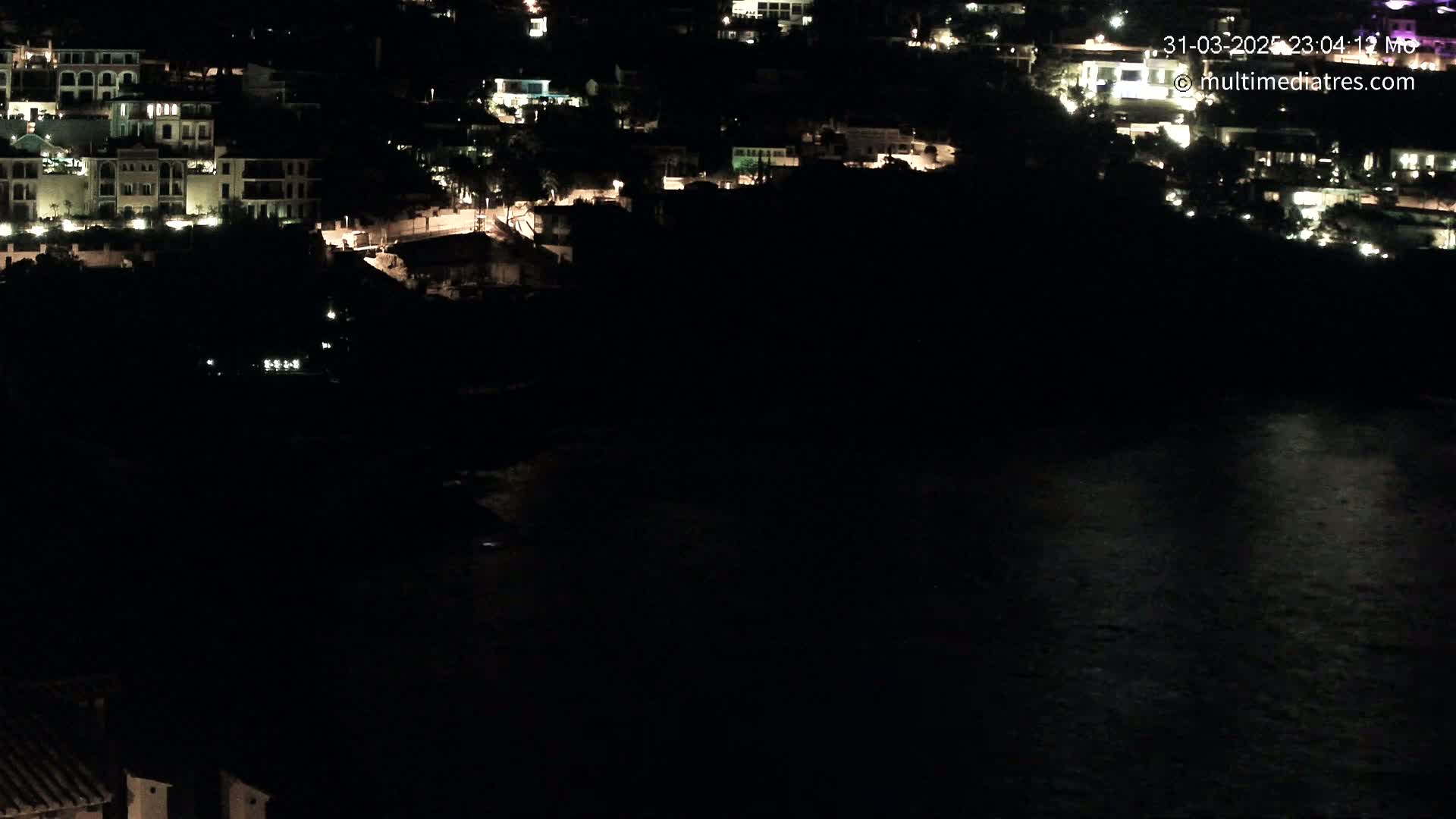 A nighttime view of a coastal town with illuminated buildings and a dark body of water reflecting some light.