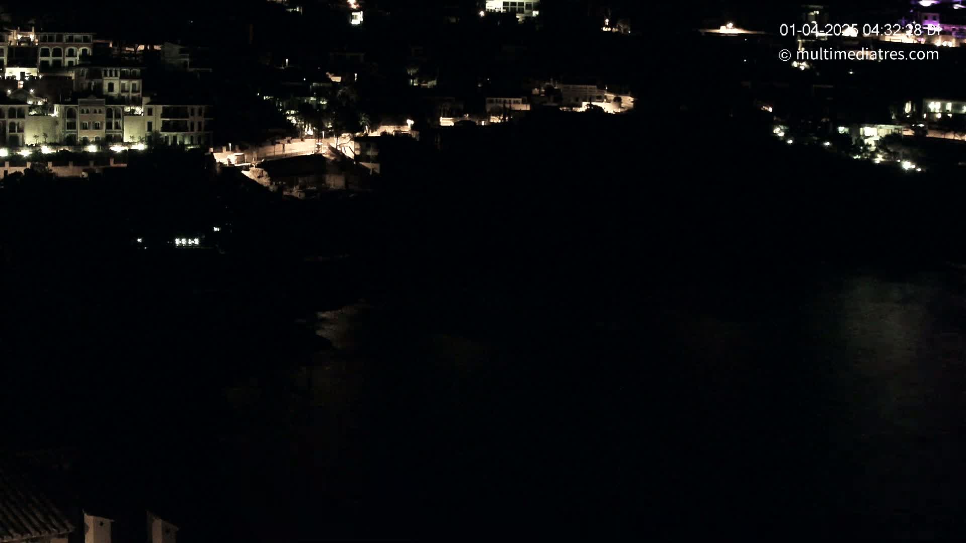 A nighttime, high-angle view shows a collection of buildings with illuminated windows near a dark body of water.
