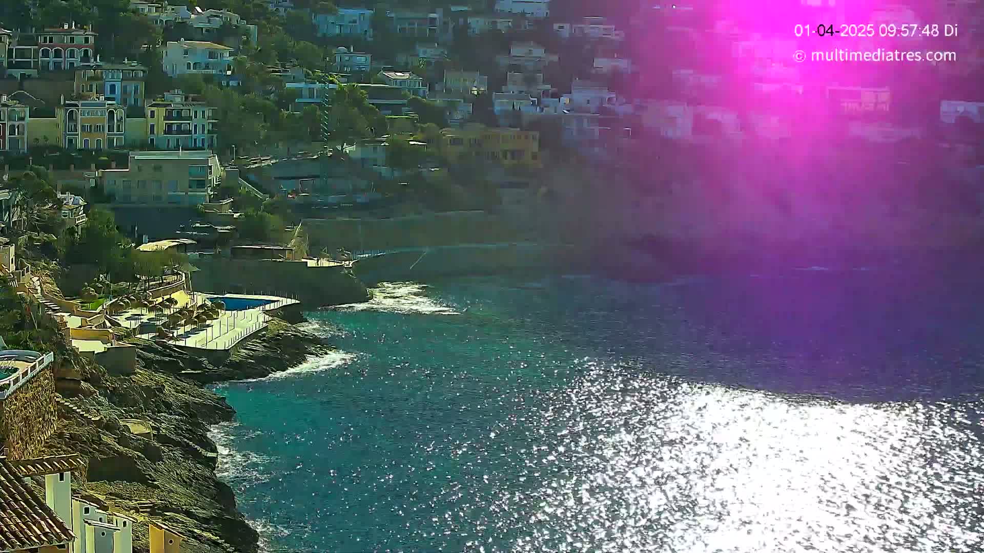 A rocky coastline with residential buildings is shown, overlooking a sparkling blue sea on a sunny day.