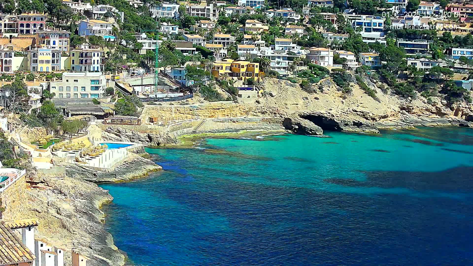 A rocky coastline with turquoise water features numerous houses perched on a hillside, under sunny conditions.